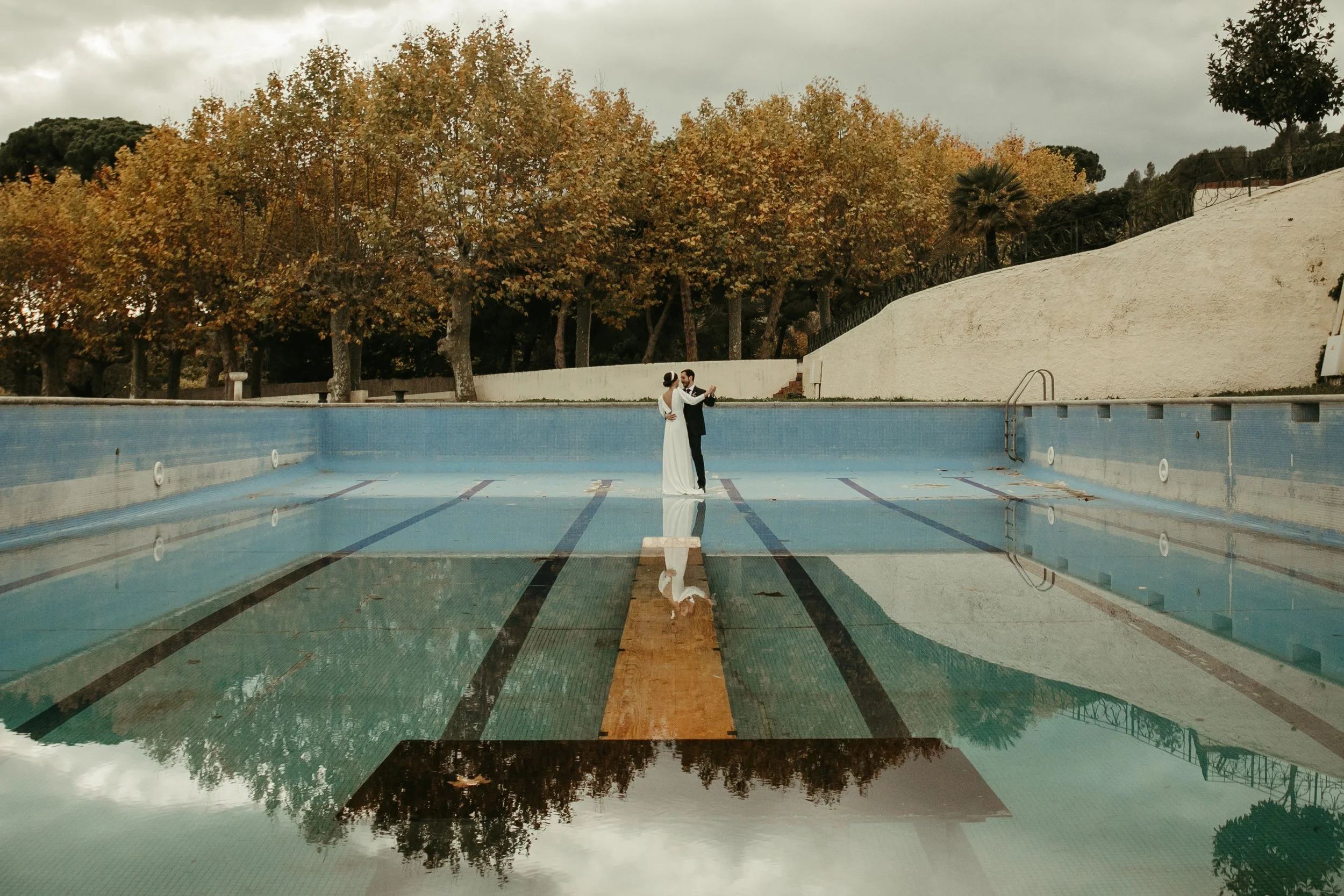 Pareja de novios bailando en una piscina vacía, con árboles de hojas otoñales al fondo.
