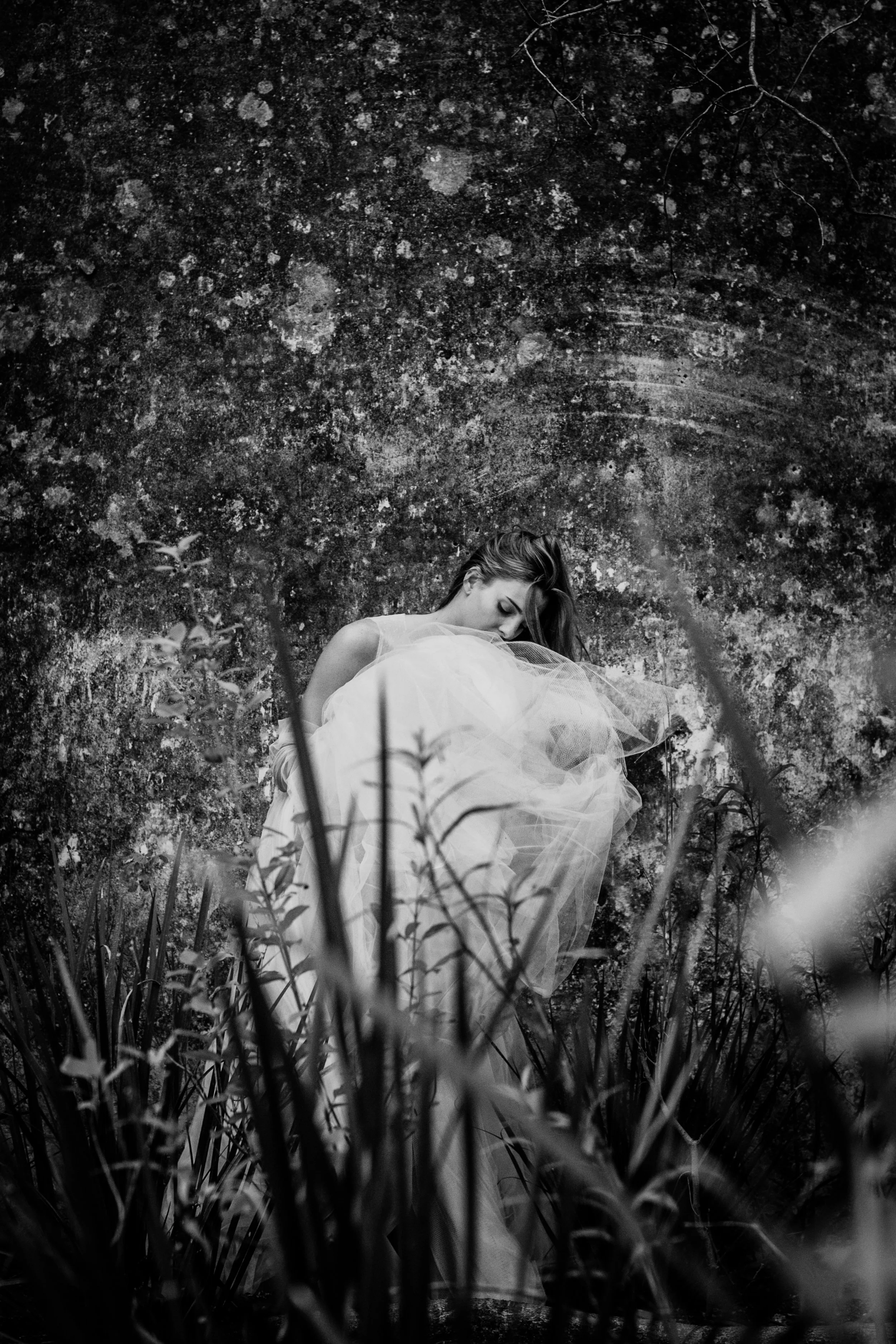 Una mujer con vestido largo y de color claro, rodeada de vegetación, de espaldas, en un entorno natural, en blanco y negro.