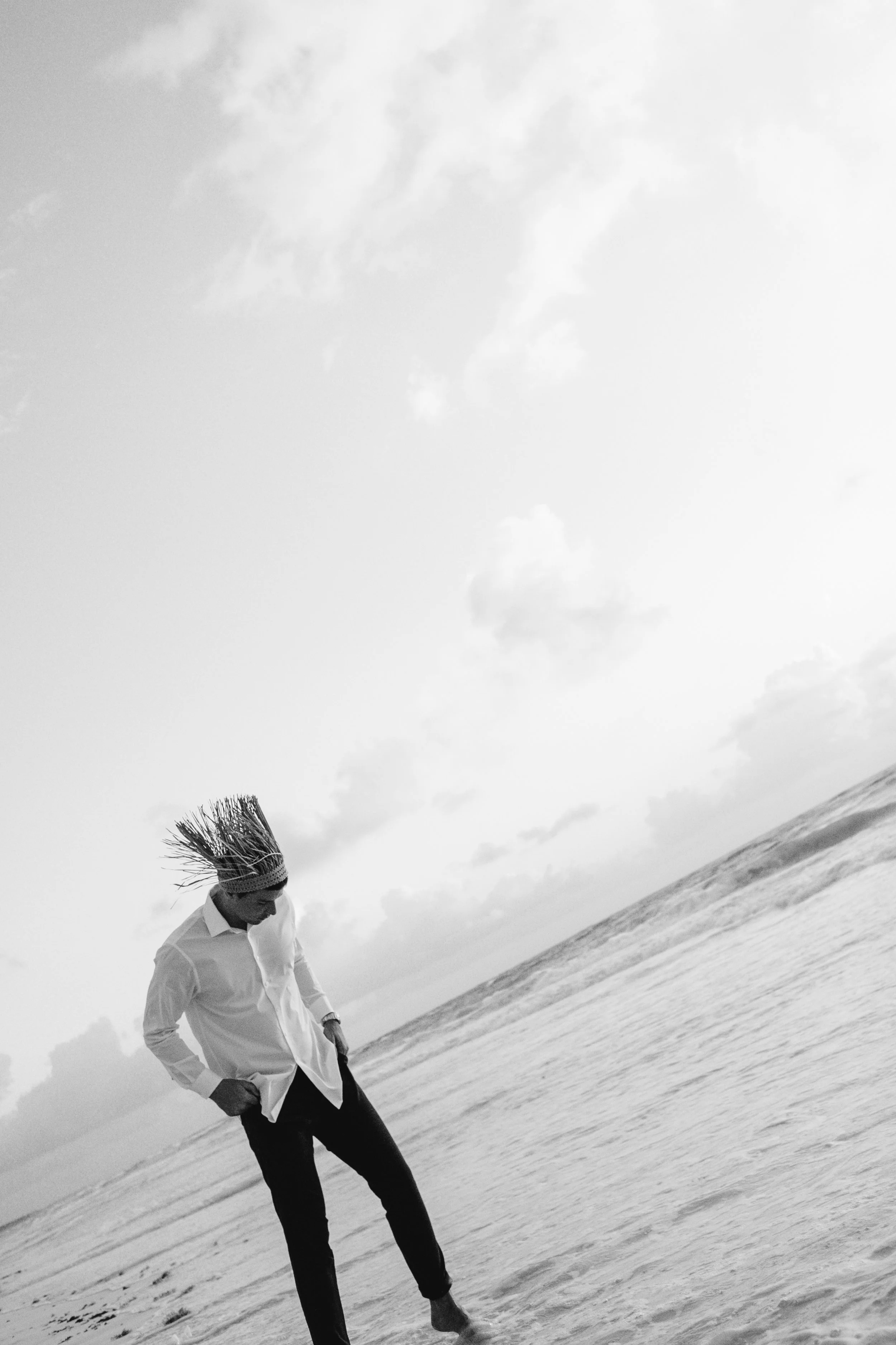 Persona con sombrero de paja, camisa blanca y pantalones negros en la orilla del mar mirando hacia abajo.