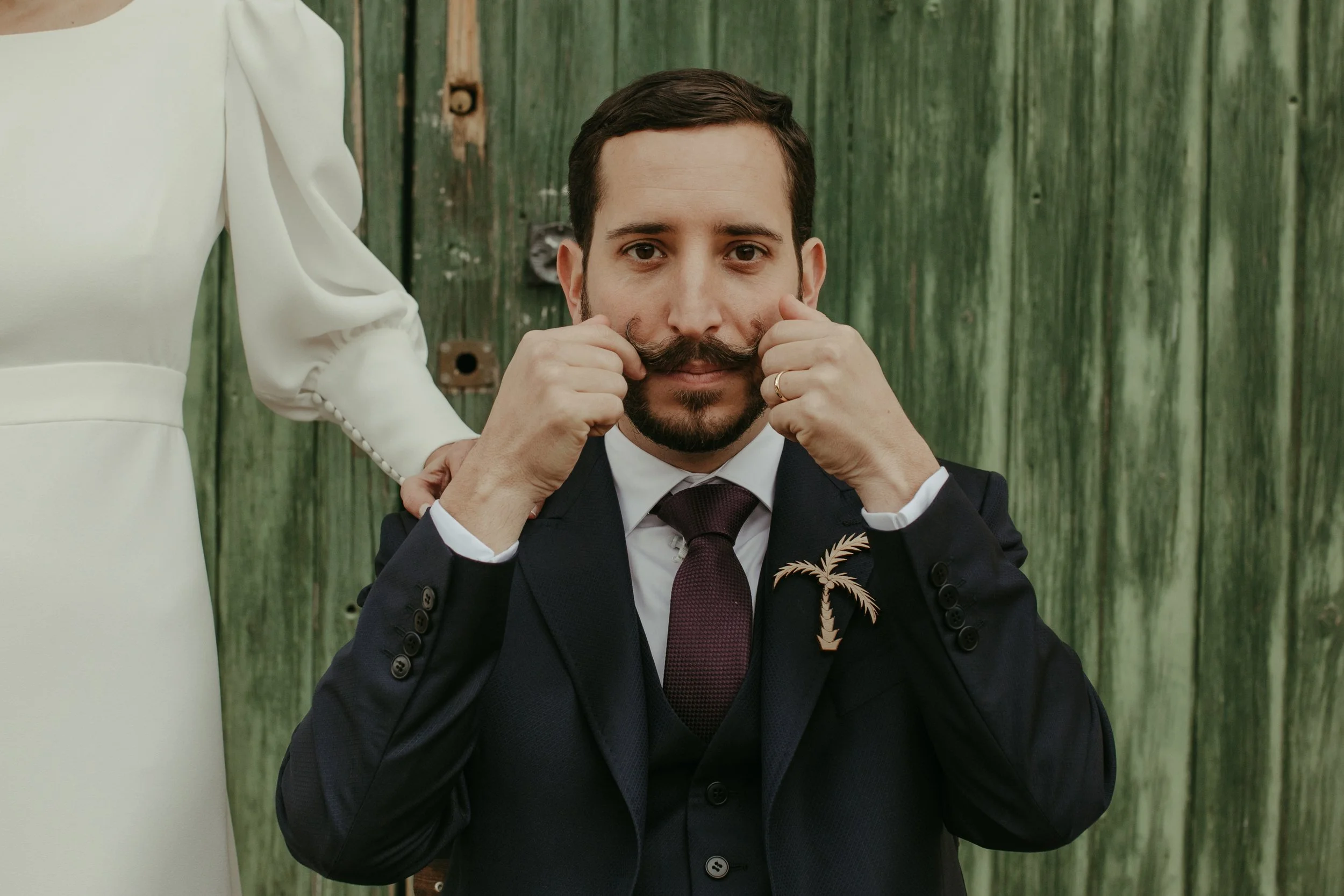 Hombre con barba y bigote, vestido con saco oscuro, camisa blanca y corbata, sin bigote, tocando sus mejillas con ambas manos, con una persona vestida de blanco detrás, con fondo de madera verde.