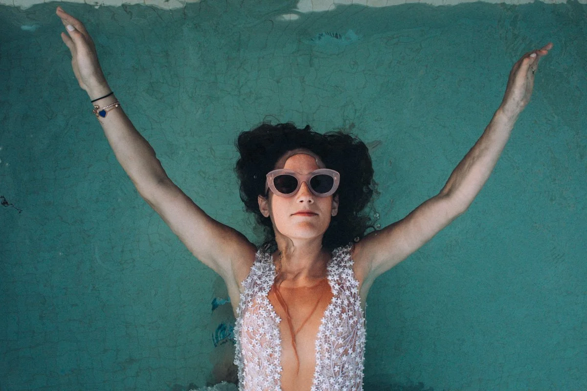 Una mujer con cabello rizado oscuro, usando gafas grandes de sol, vestida con un traje de baño con encaje y detalles de flores, está flotando en una piscina con los brazos extendidos hacia los lados.