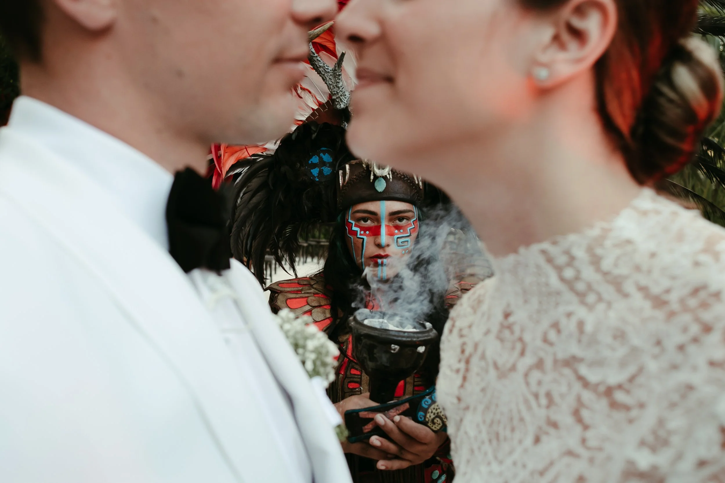 Una pareja en su boda se acerca para darse un beso, mientras una mujer con vestimenta tradicional indígena y pintada facialmente observa en el fondo, con una caldera humeante en las manos.