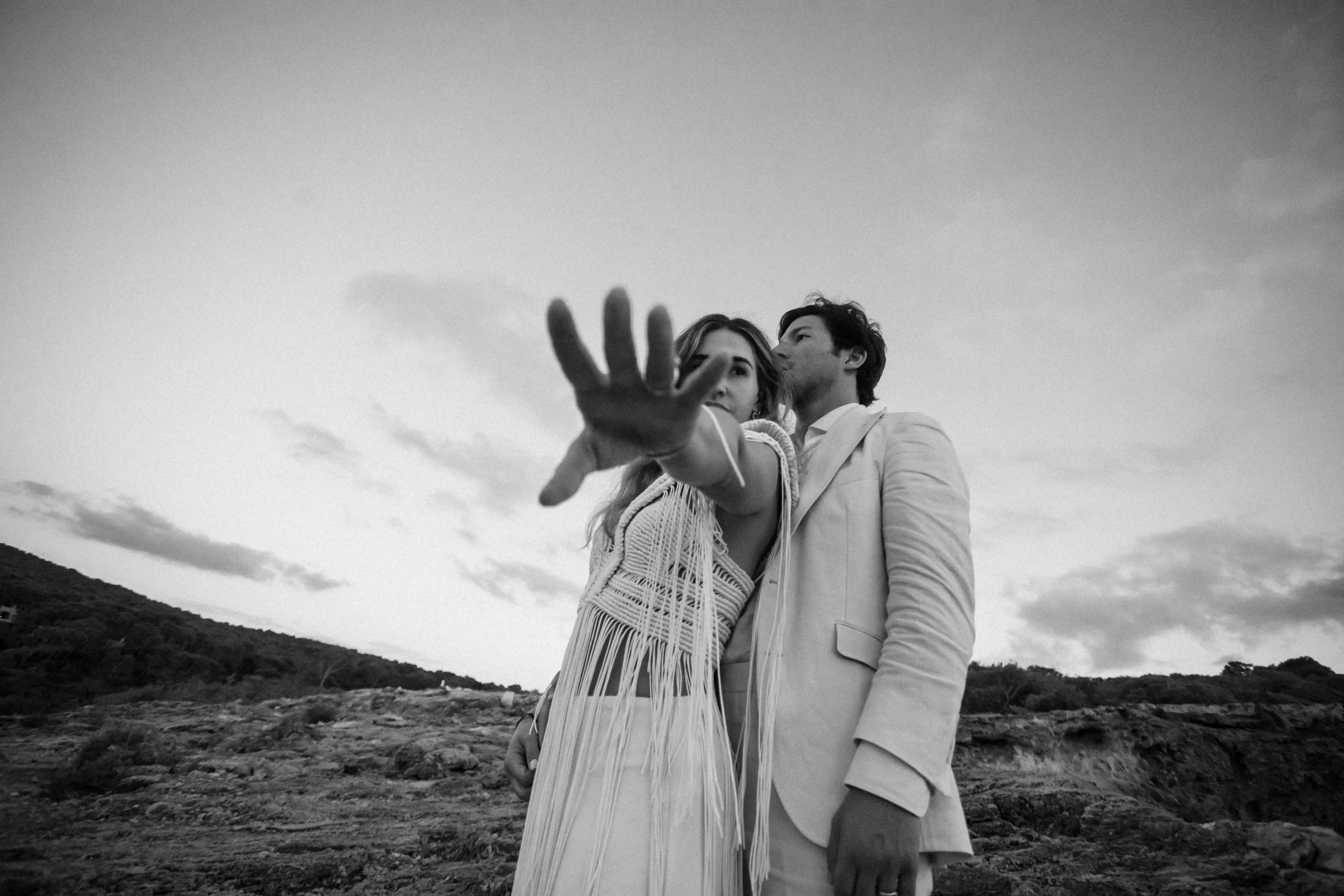 Una pareja en una escenografía natural, con fondo de cielo y montañas, uno de ellos extendiendo la mano hacia la cámara y ella mirándolo mientras él la besa en la frente, en blanco y negro.