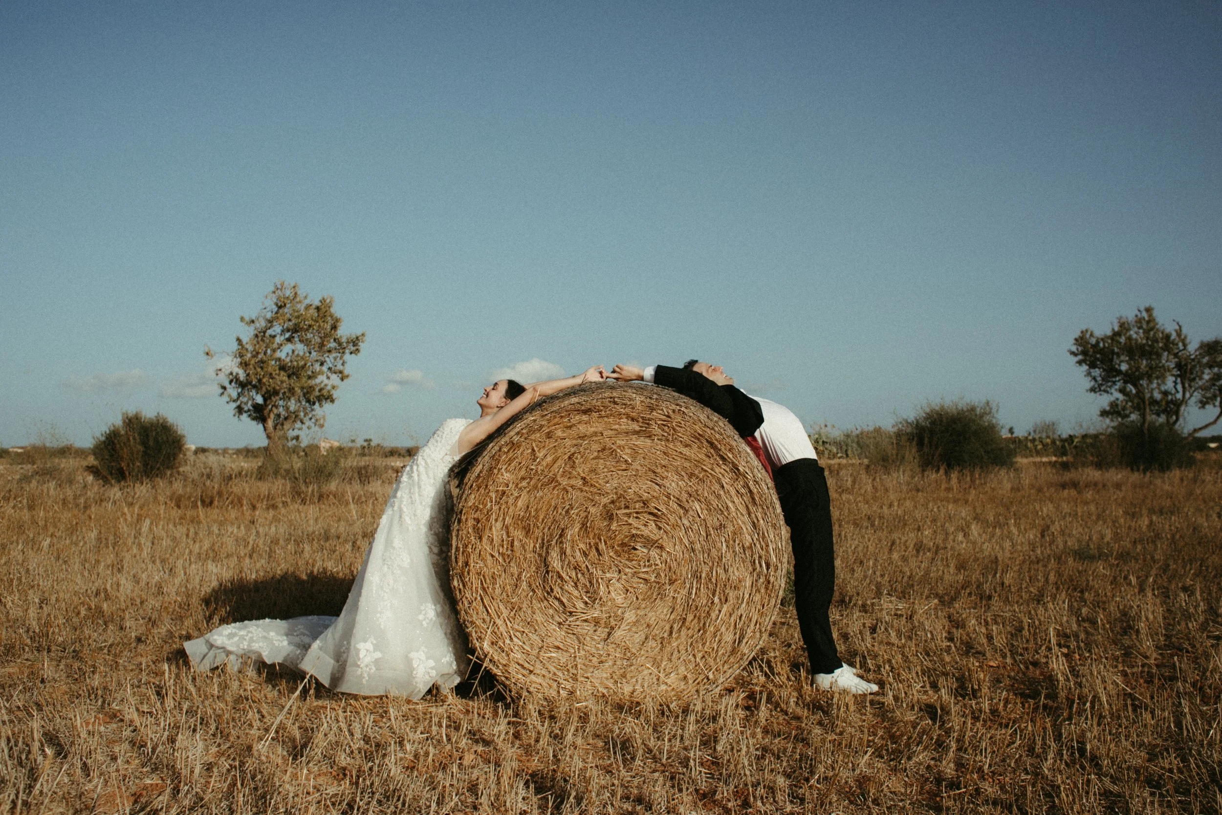 Una pareja de novios en un campo abierto con un gran tambor de heno entre ellos, con árboles dispersos y un cielo despejado.