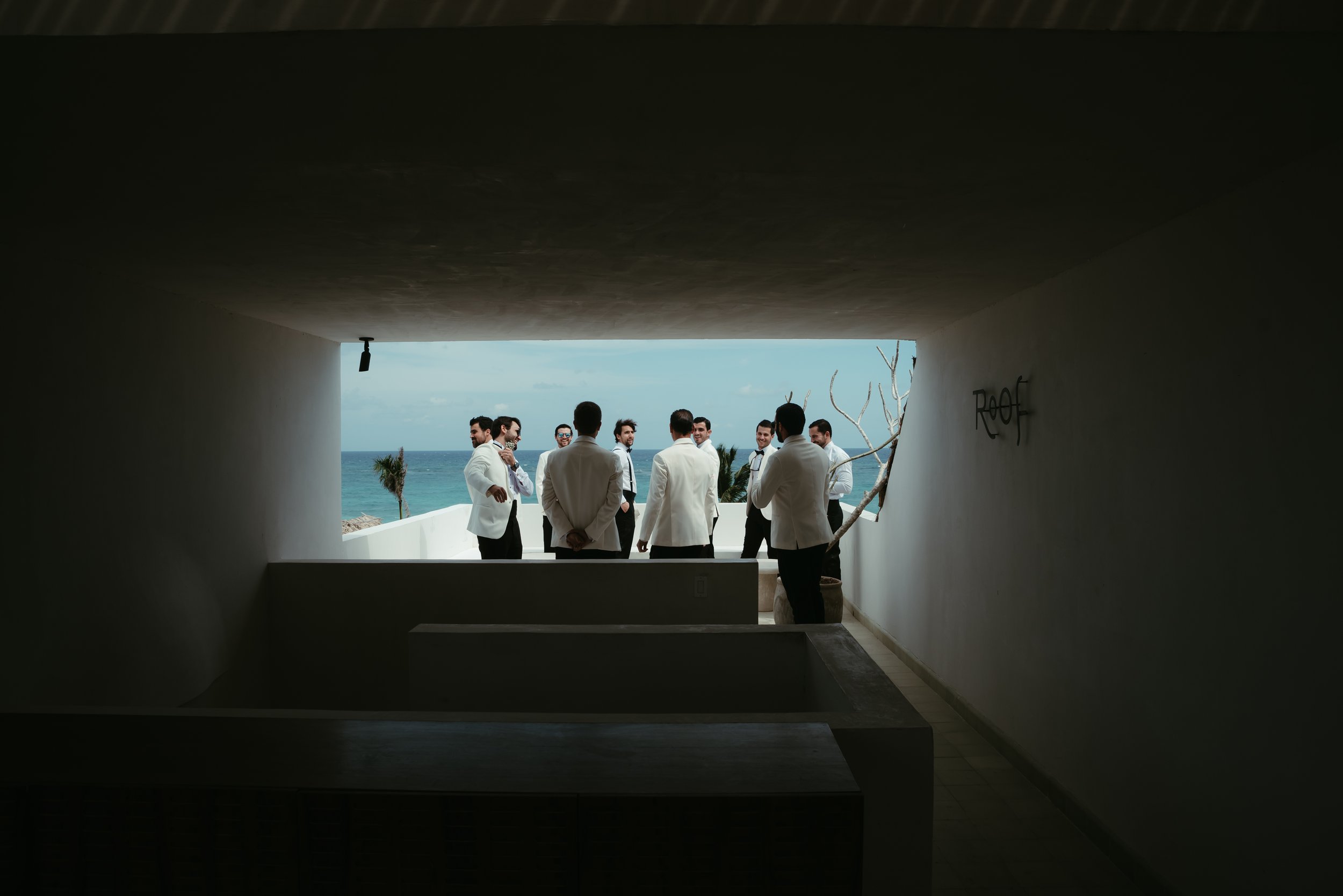 Grupo de hombres con trajes blancos y negros en una terraza con vista al mar, vista desde un espacio sombreado.