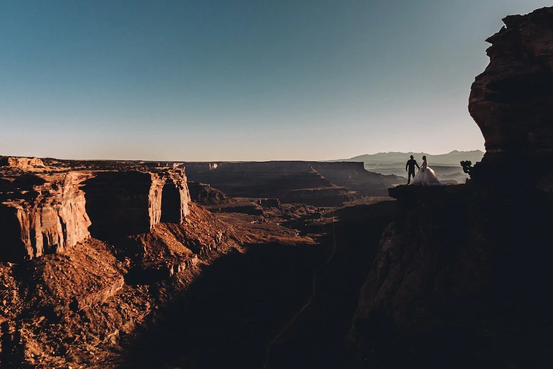 Una pareja camina por un acantilado en un paisaje desértico durante el atardecer, con formaciones rocosas y un cielo despejado.