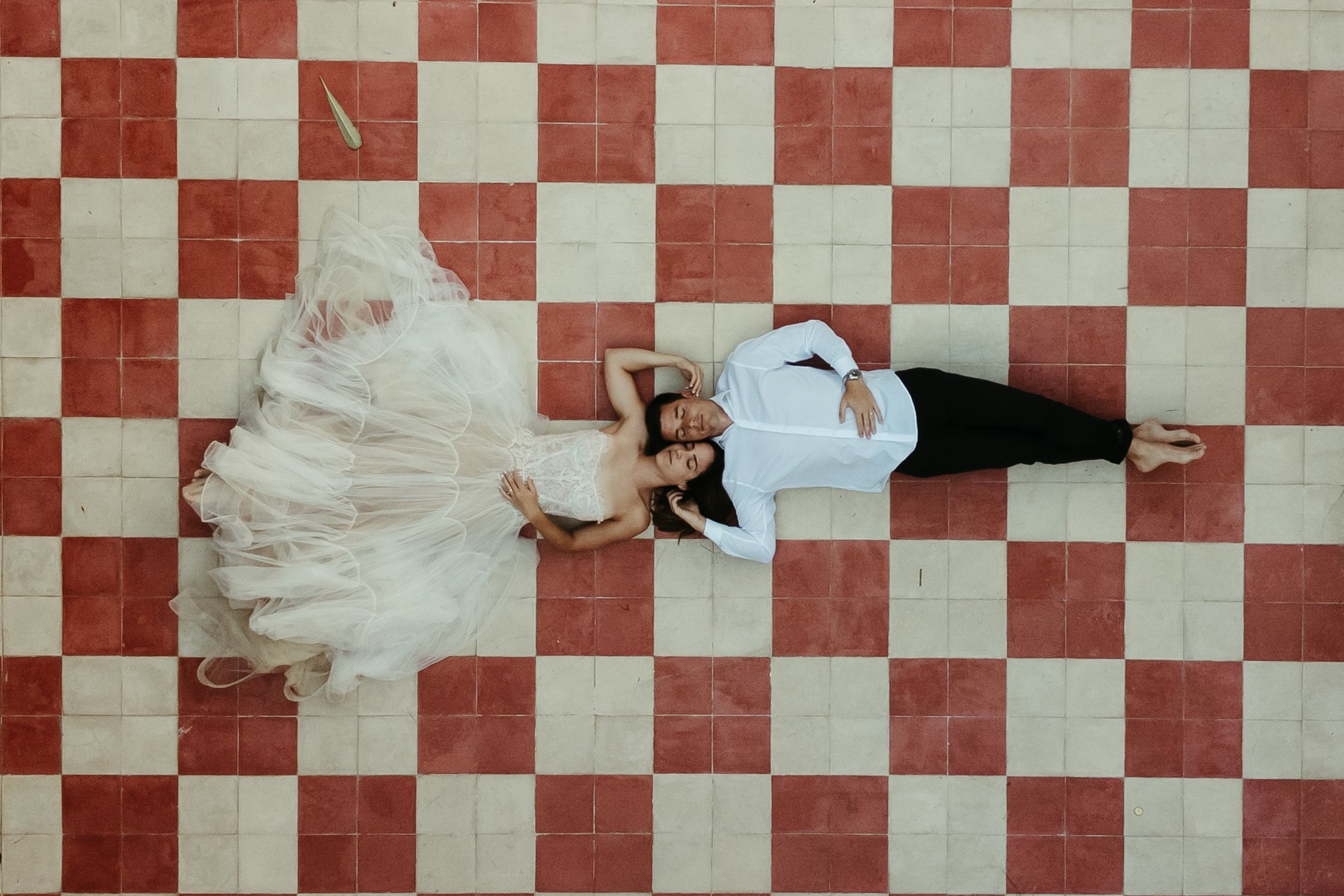 Pareja acostada sobre un piso de baldosas rojas y blancas, la mujer usa vestido de novia y el hombre ropa formal blanca y negra.