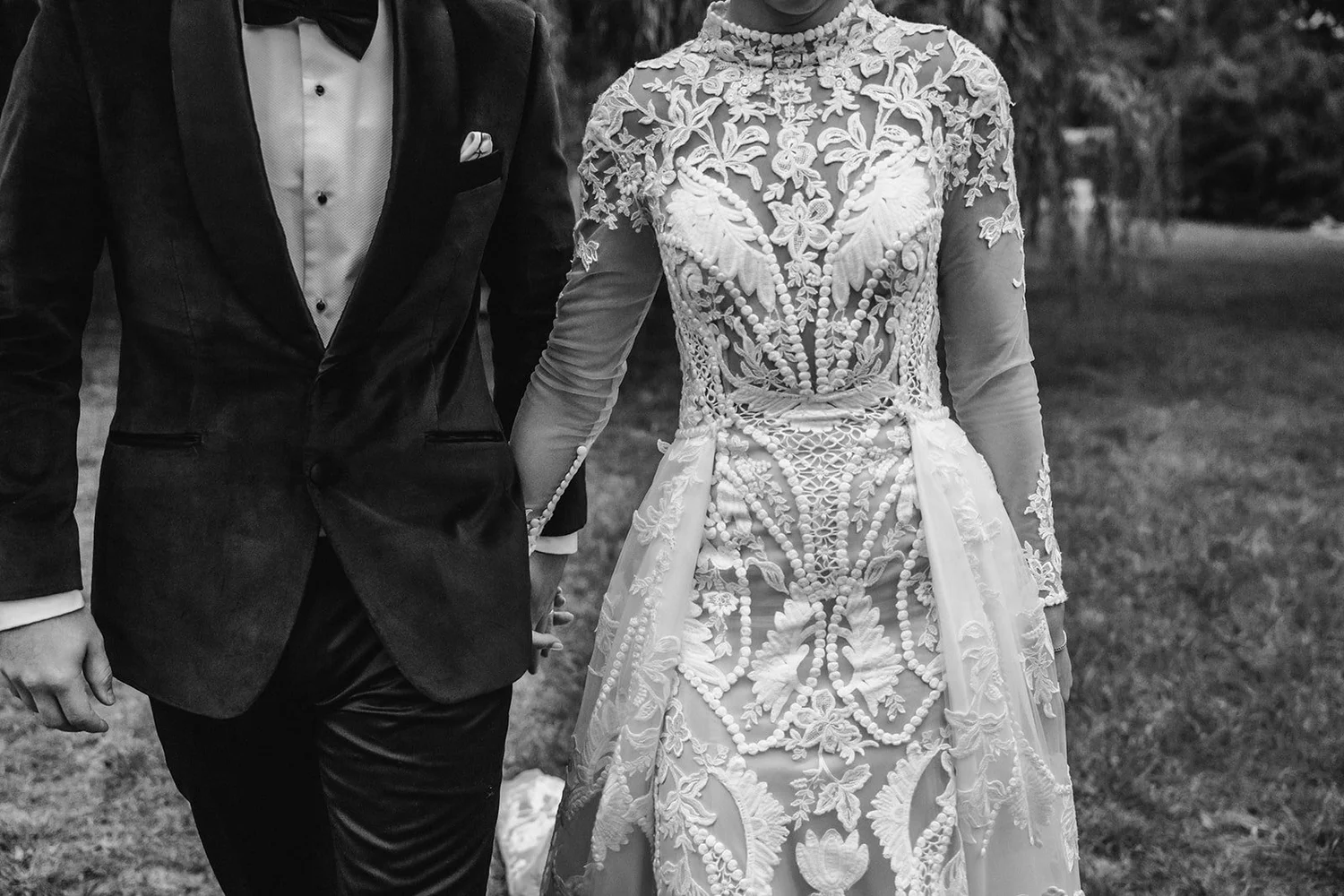 Black and white detail of the groom in a velvet tuxedo walking hand in hand with the bride in an embroidered lace dress, El Escorial