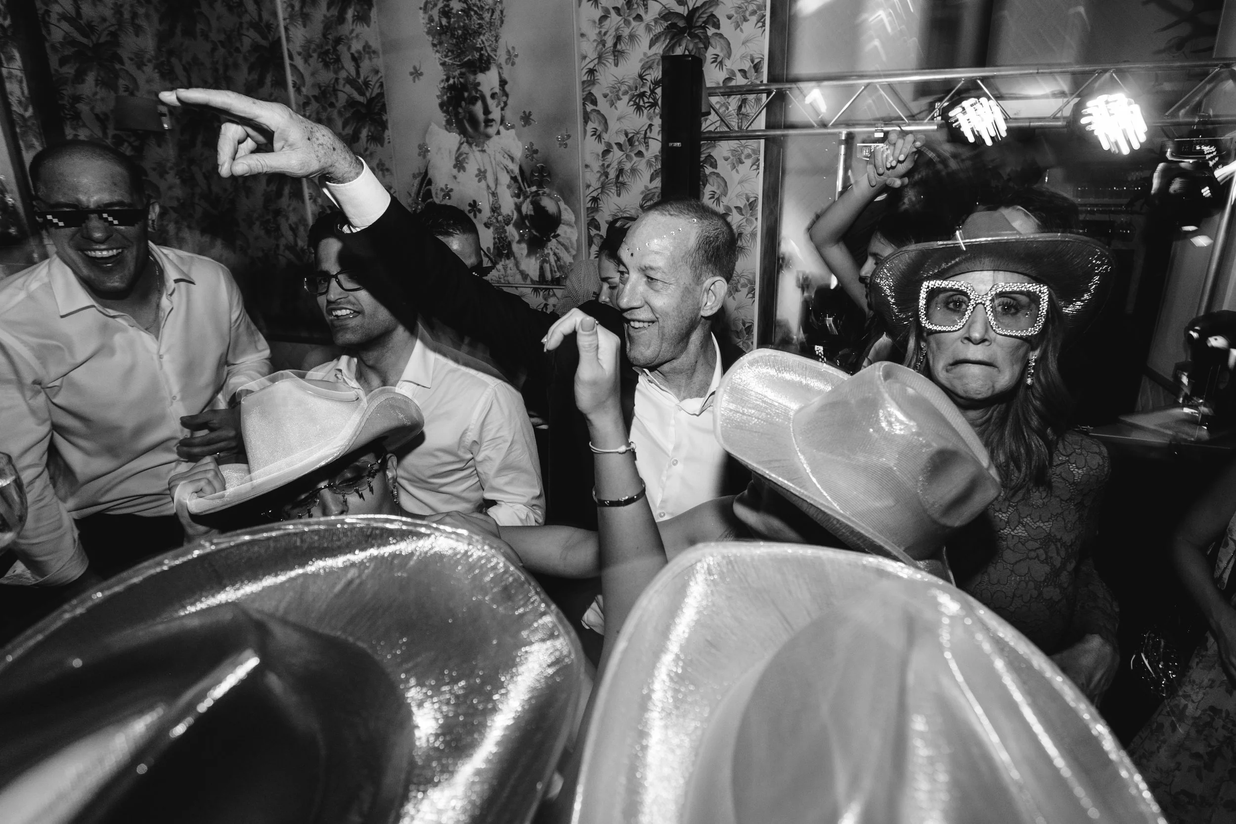 Personas en una fiesta, algunos con sombreros grandes y gafas llamativas, bailando y sonriendo en un ambiente festivo.