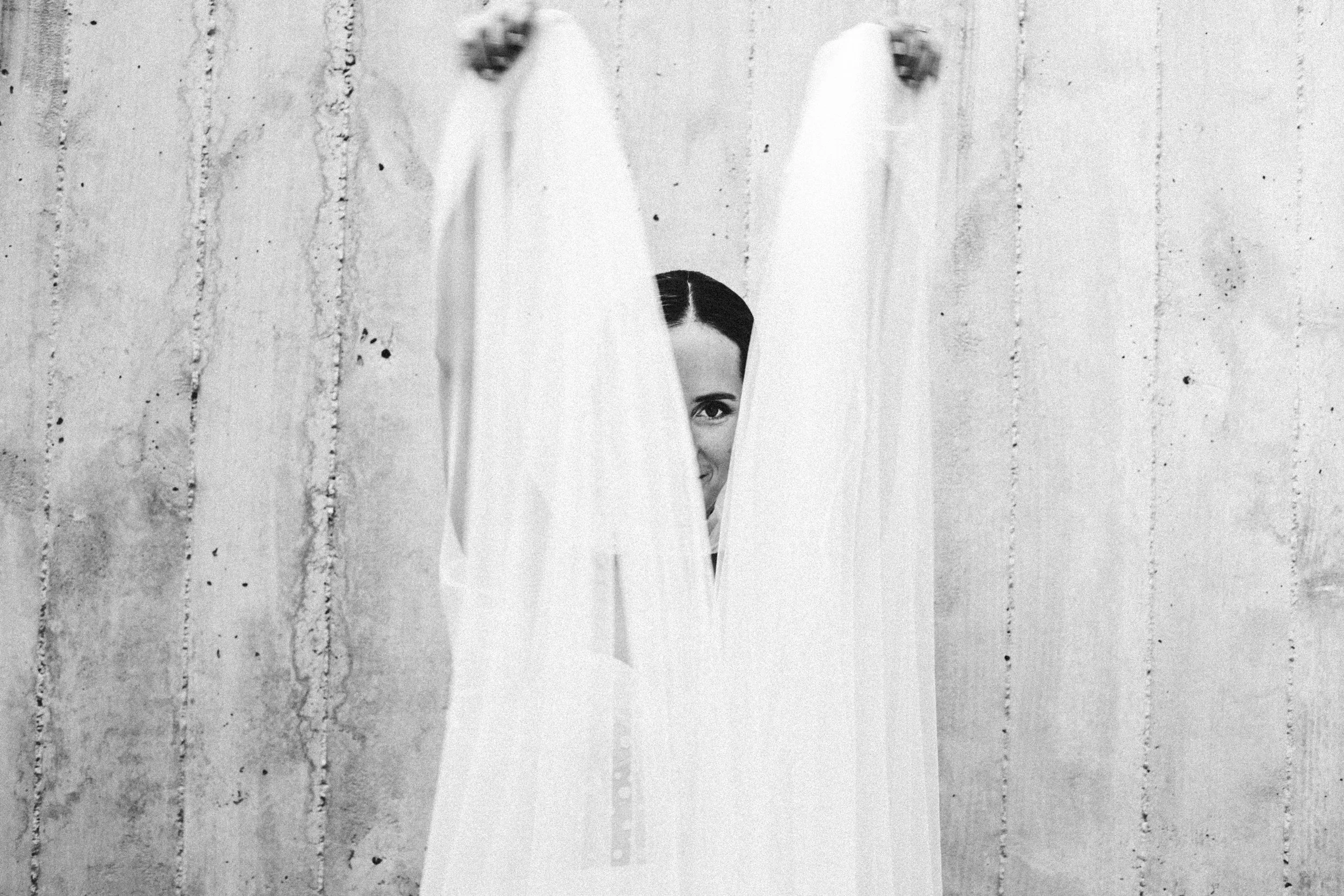 Una mujer mira a través de una abertura entre dos cortinas blancas, con una pared de fondo de aspecto rustic. Solo se ve su rostro parcialmente, con una expresión sonriente.