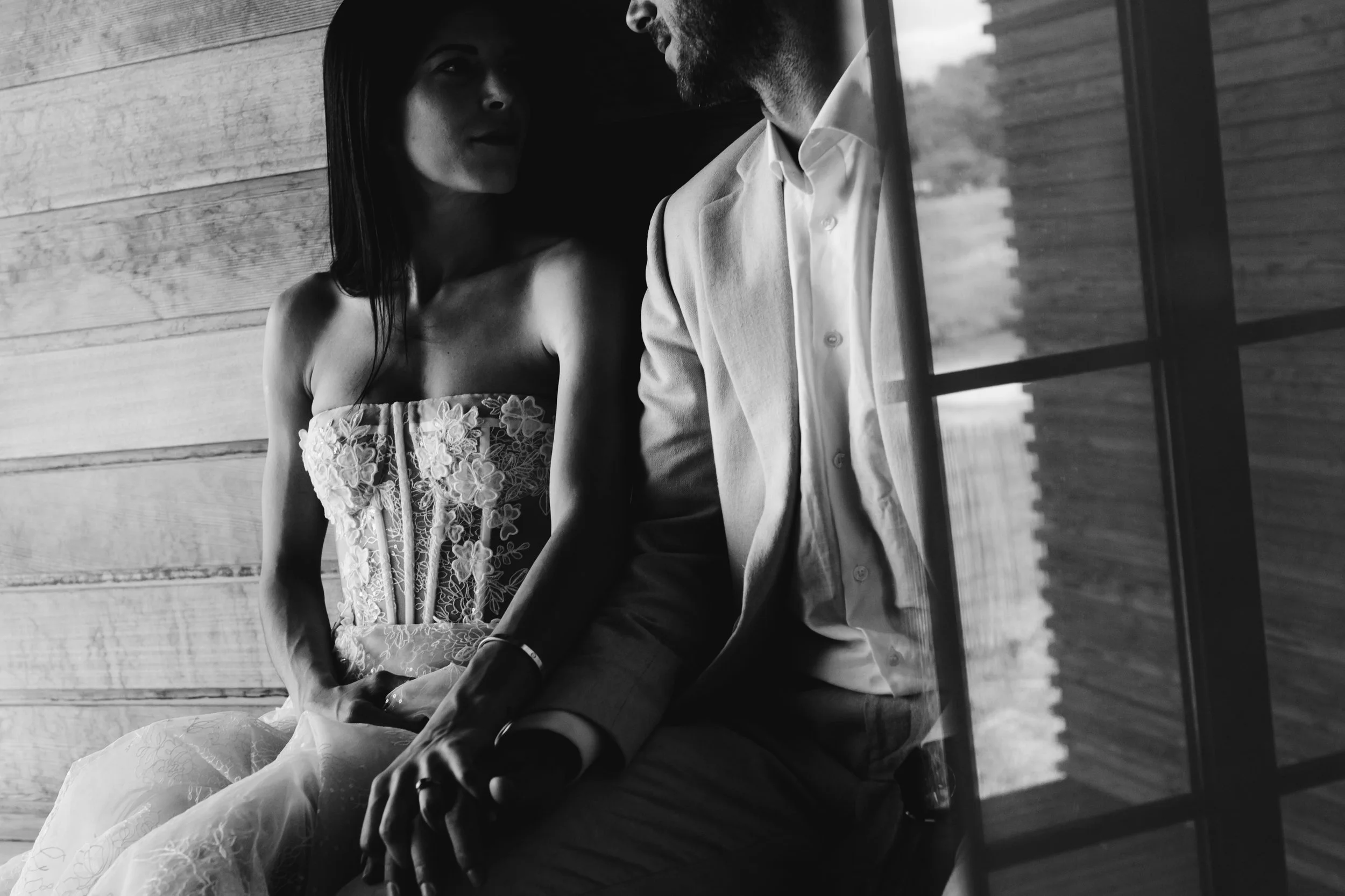 Una pareja sentada cerca de una ventana de madera, en un ambiente tranquilo, la mujer lleva un vestido sin tirantes con detalles de encaje y el hombre viste una chaqueta y una camisa. La imagen es en blanco y negro.