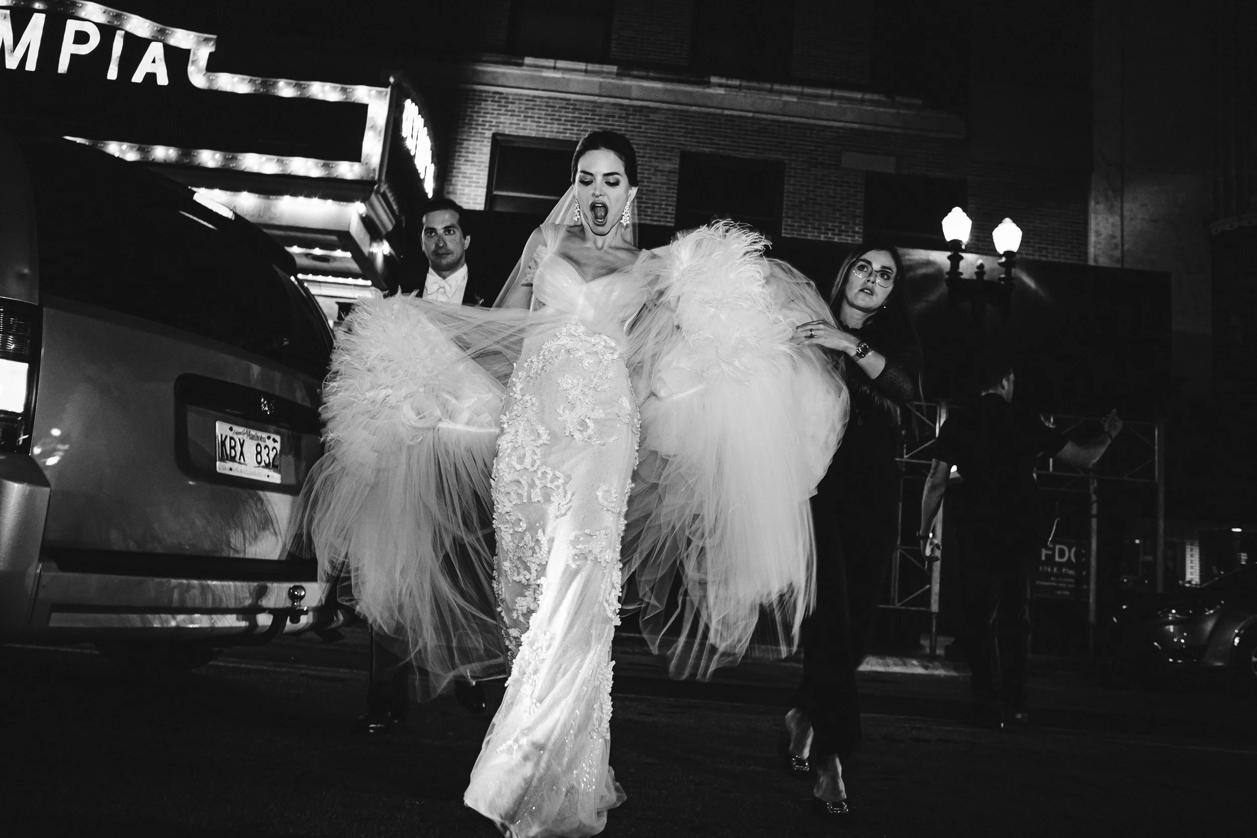 Una mujer con un vestido de novia brillante y un pañuelo en la cabeza sale de un auto en la calle por la noche, rodeada de personas.