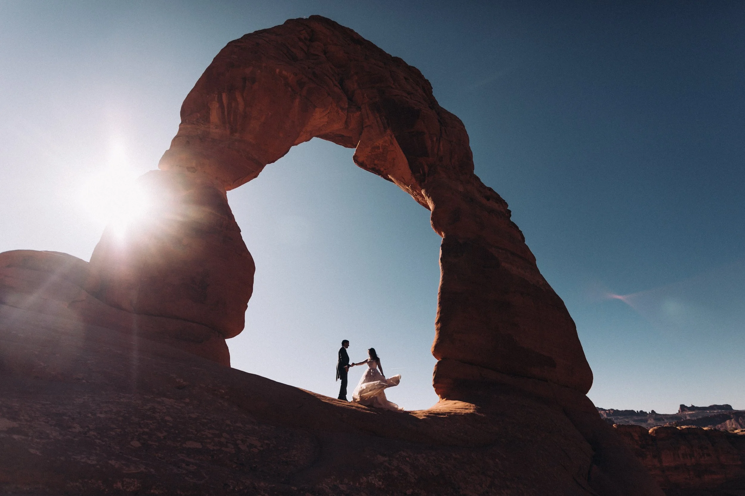 Pareja en vestido de novia y traje en un arco de piedra natural en un paisaje desértico con cielo despejado y sol brillante.