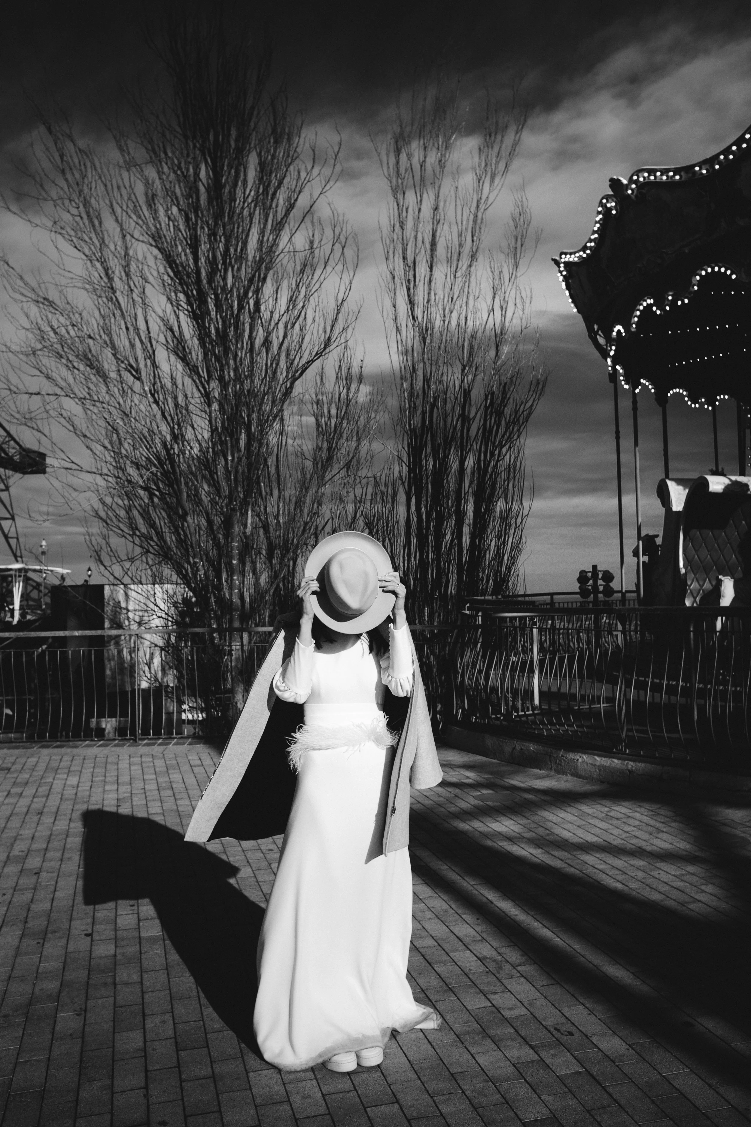 Mujer vestida con vestido largo y abrigo, sosteniendo un sombrero y cubriéndose la cara, en un parque con carrusel y árboles sin hojas, en una escena en blanco y negro.
