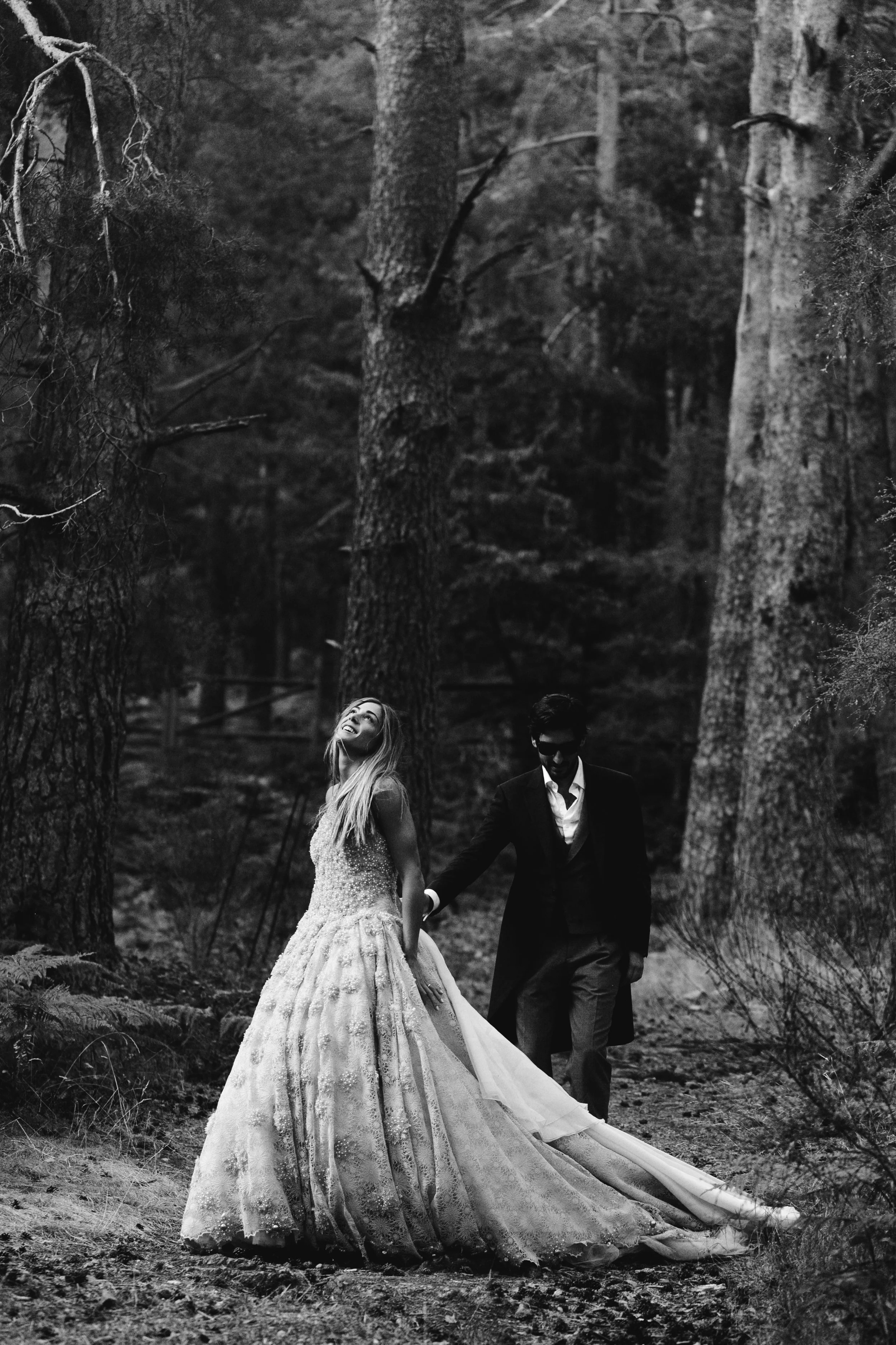 Una pareja vestida con ropa de boda en un bosque, la mujer de vestido largo y el hombre con traje oscuro, en la imagen en blanco y negro.