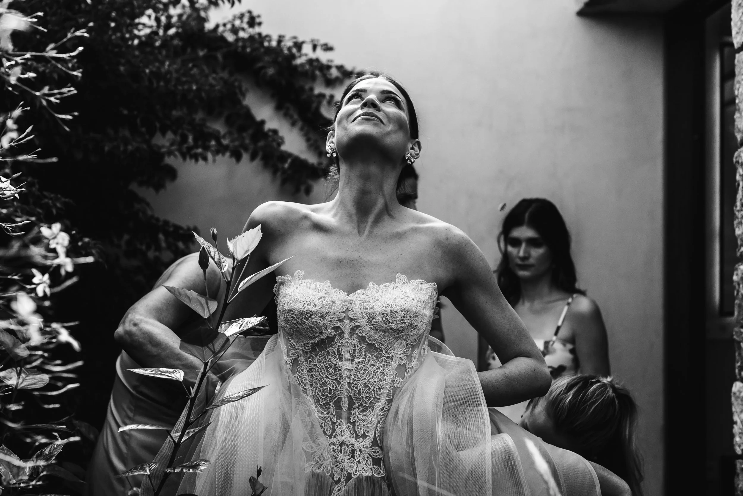 Mujer en vestido de novia con expresión de alegría, rodeada de otras personas, en un entorno exterior con plantas y paredes.