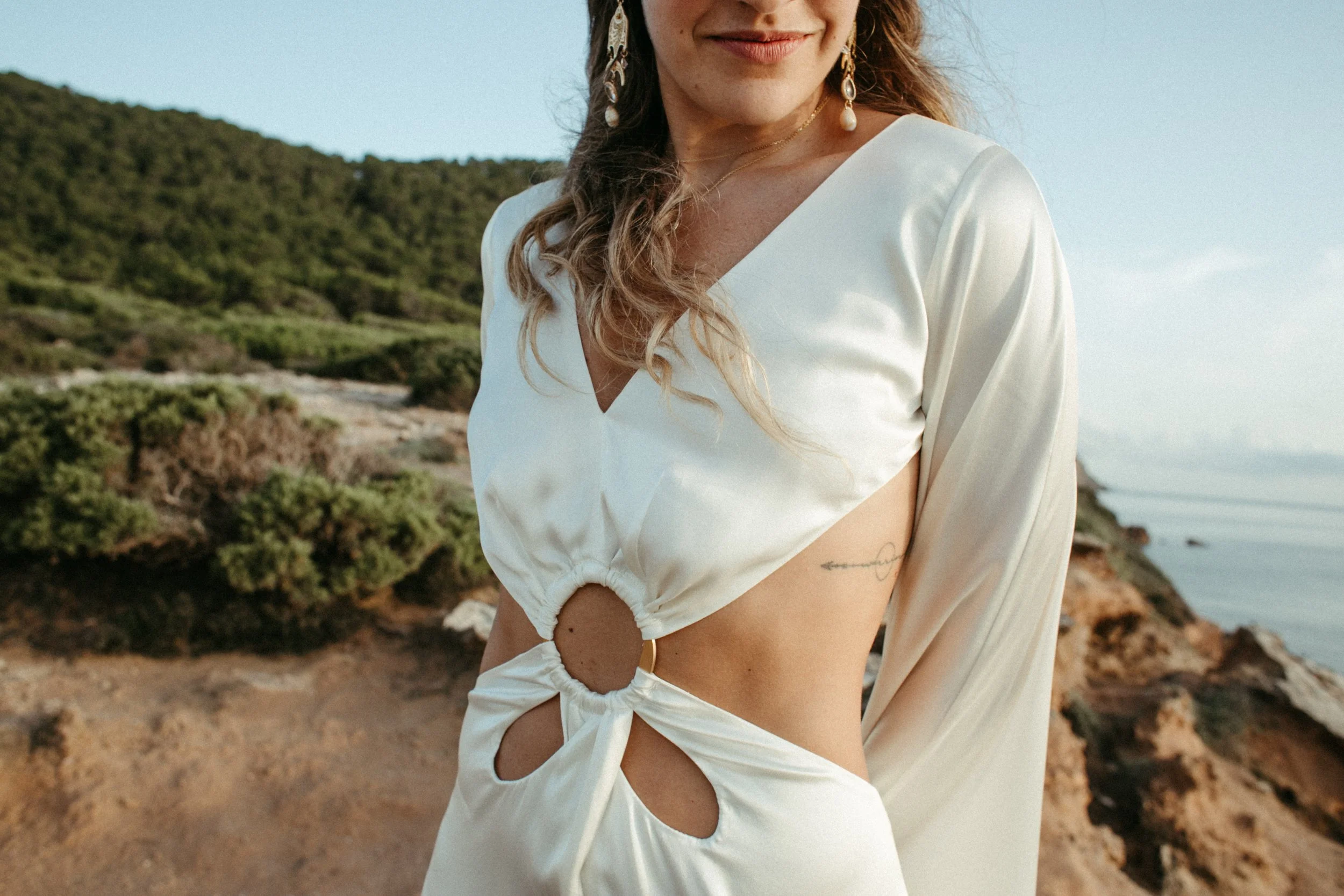 Mujer con vestido blanco con aberturas y anillo en la cintura, en un paisaje de costa con vegetación y mar al fondo.