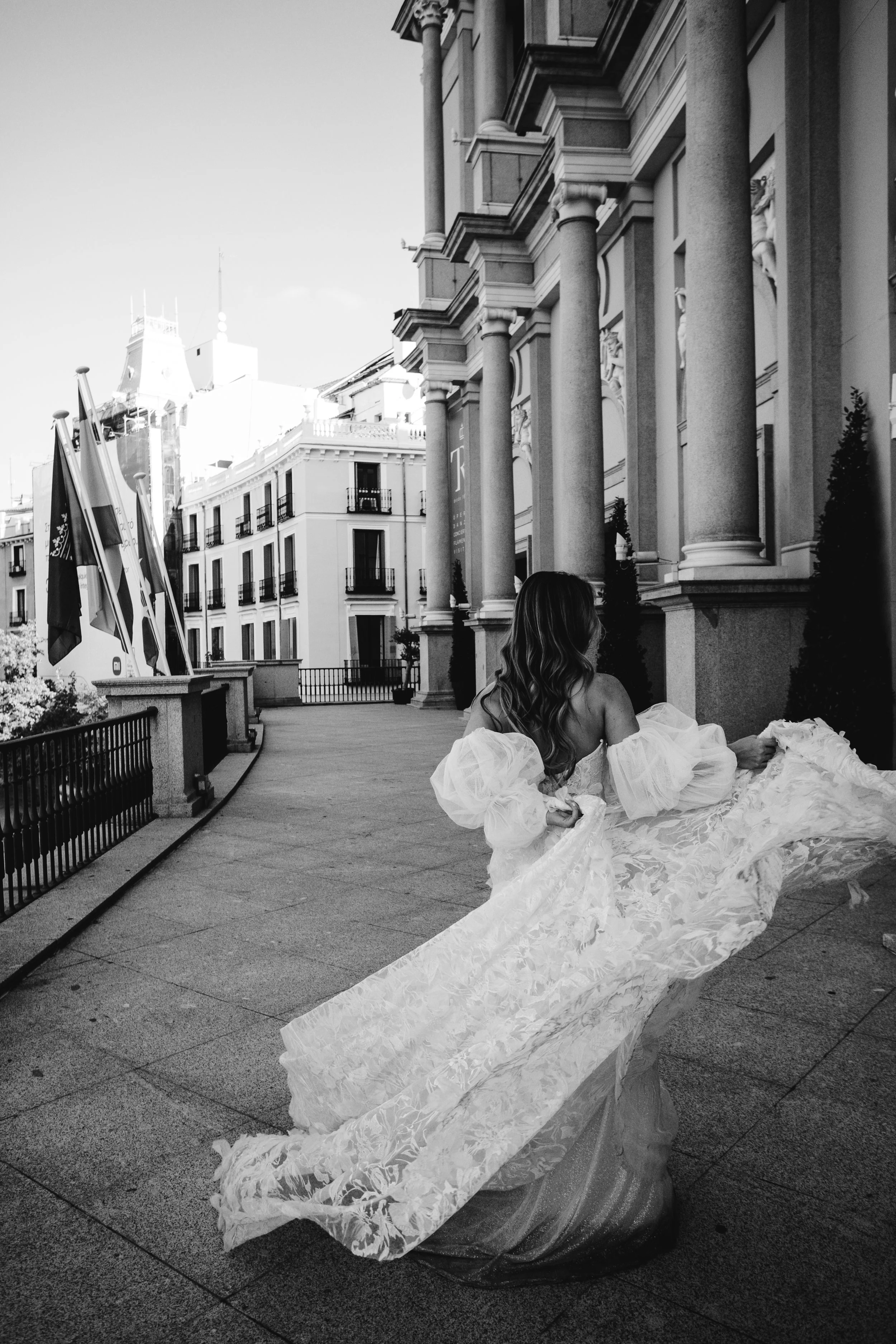 Mujer con vestido de novia de espaldas, en la calle, con edificios y columnas arquitectónicas alrededor, en una fotografía en blanco y negro.