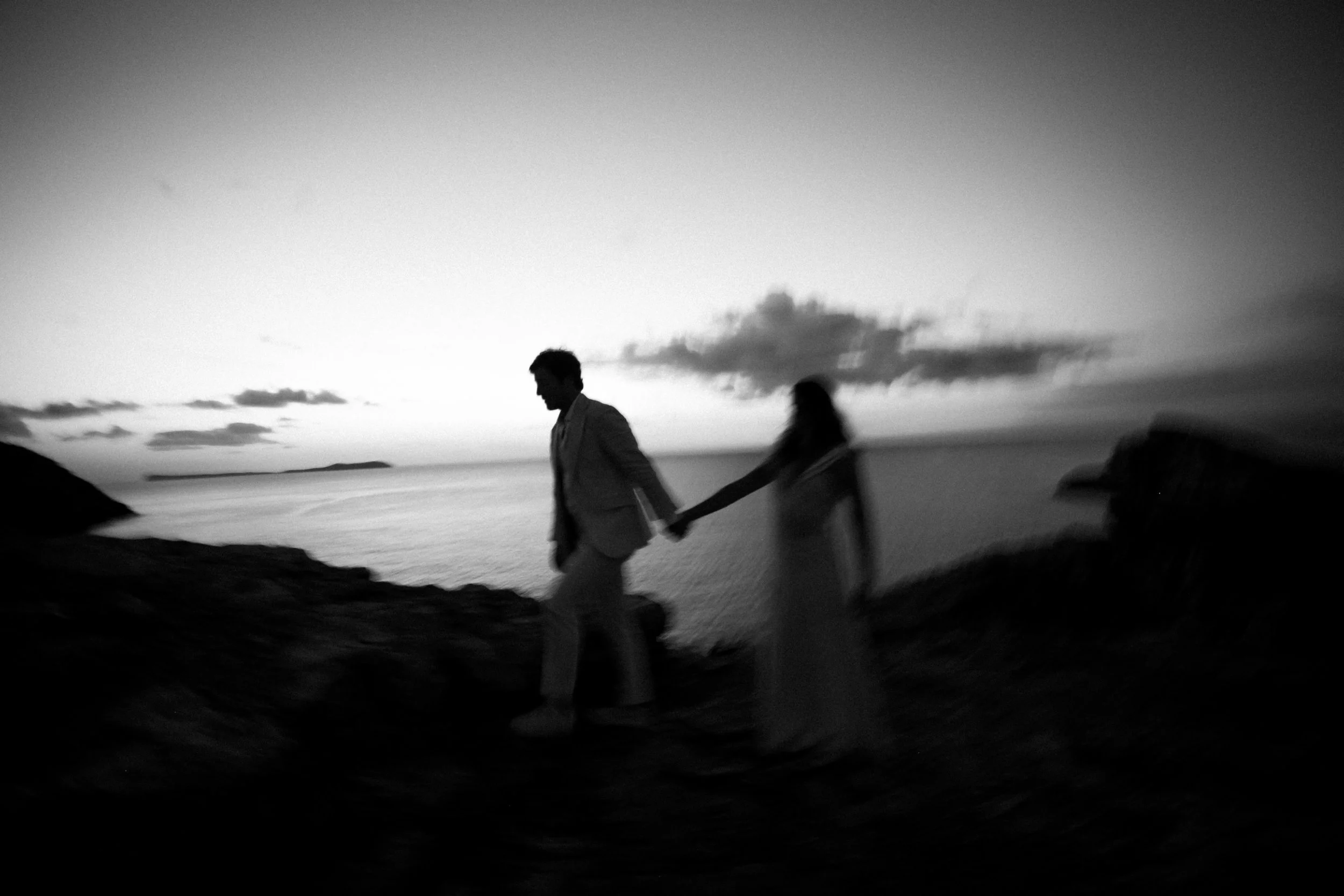 Pareja de espaldas caminando por una costa durante el atardecer, sosteniéndose de la mano, en una foto en blanco y negro.
