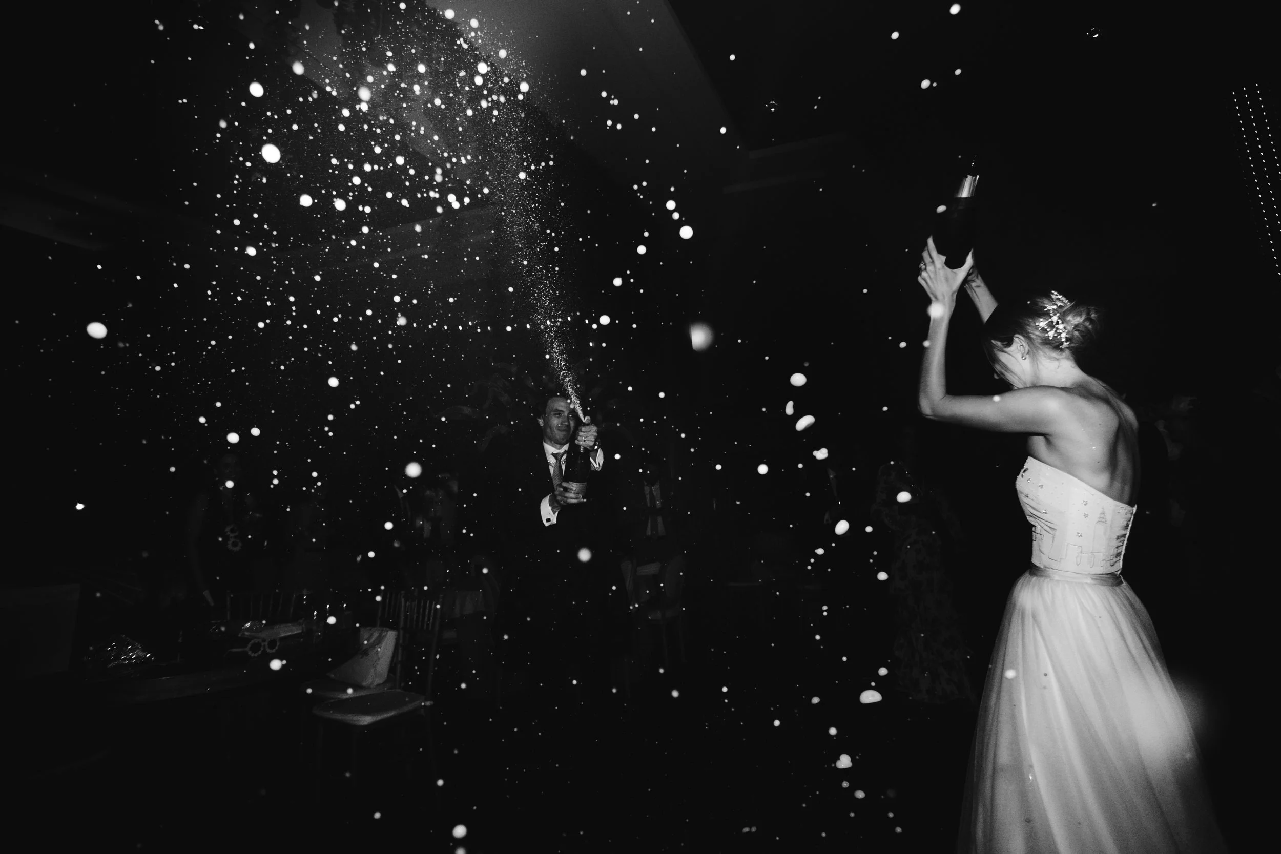 Una pareja celebrando en una boda, lanzando champagne en un ambiente oscuro, en blanco y negro.
