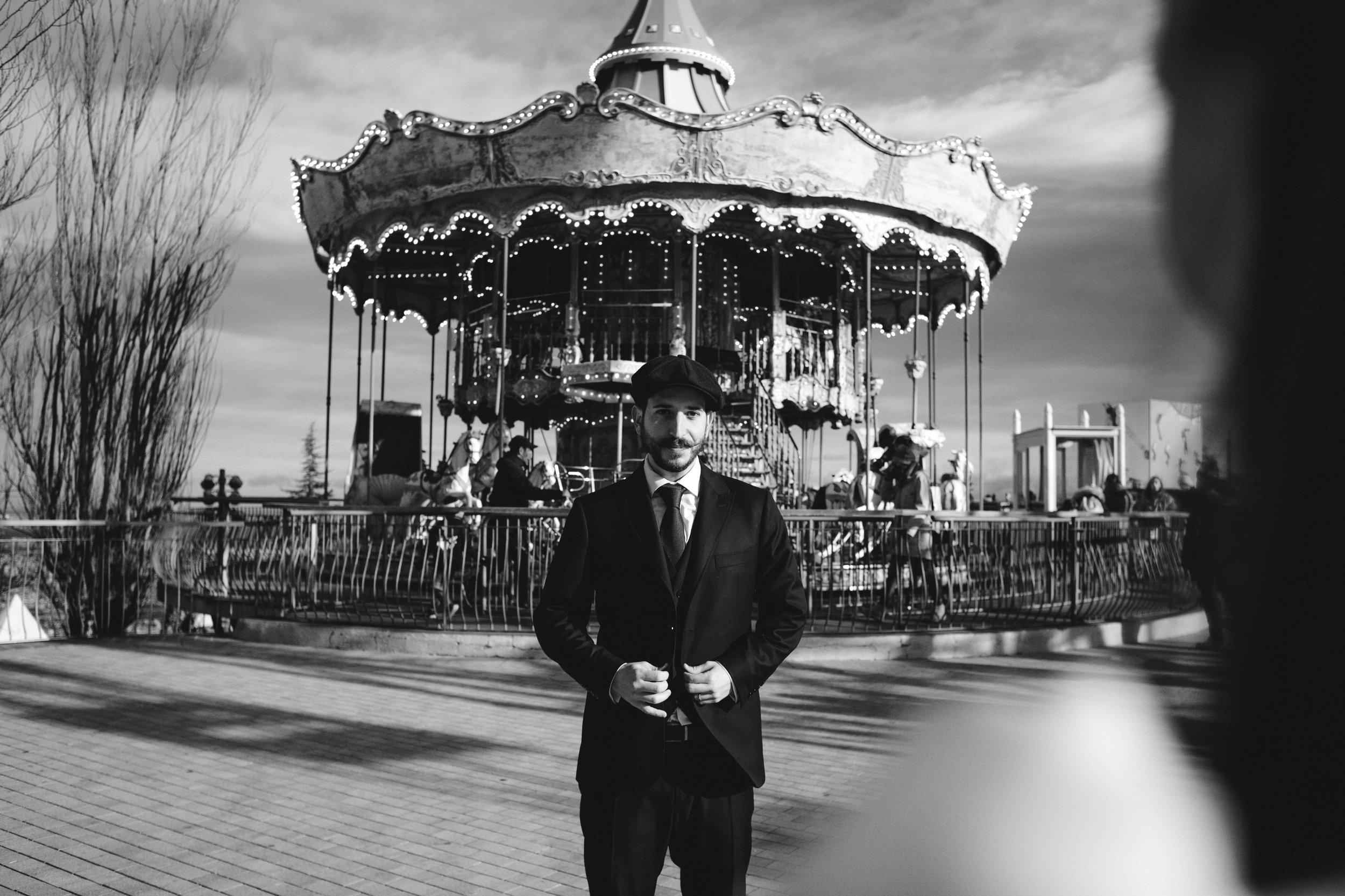 Un hombre con traje y boina posa frente a una rueda de la fortuna en un parque de diversiones al atardecer.