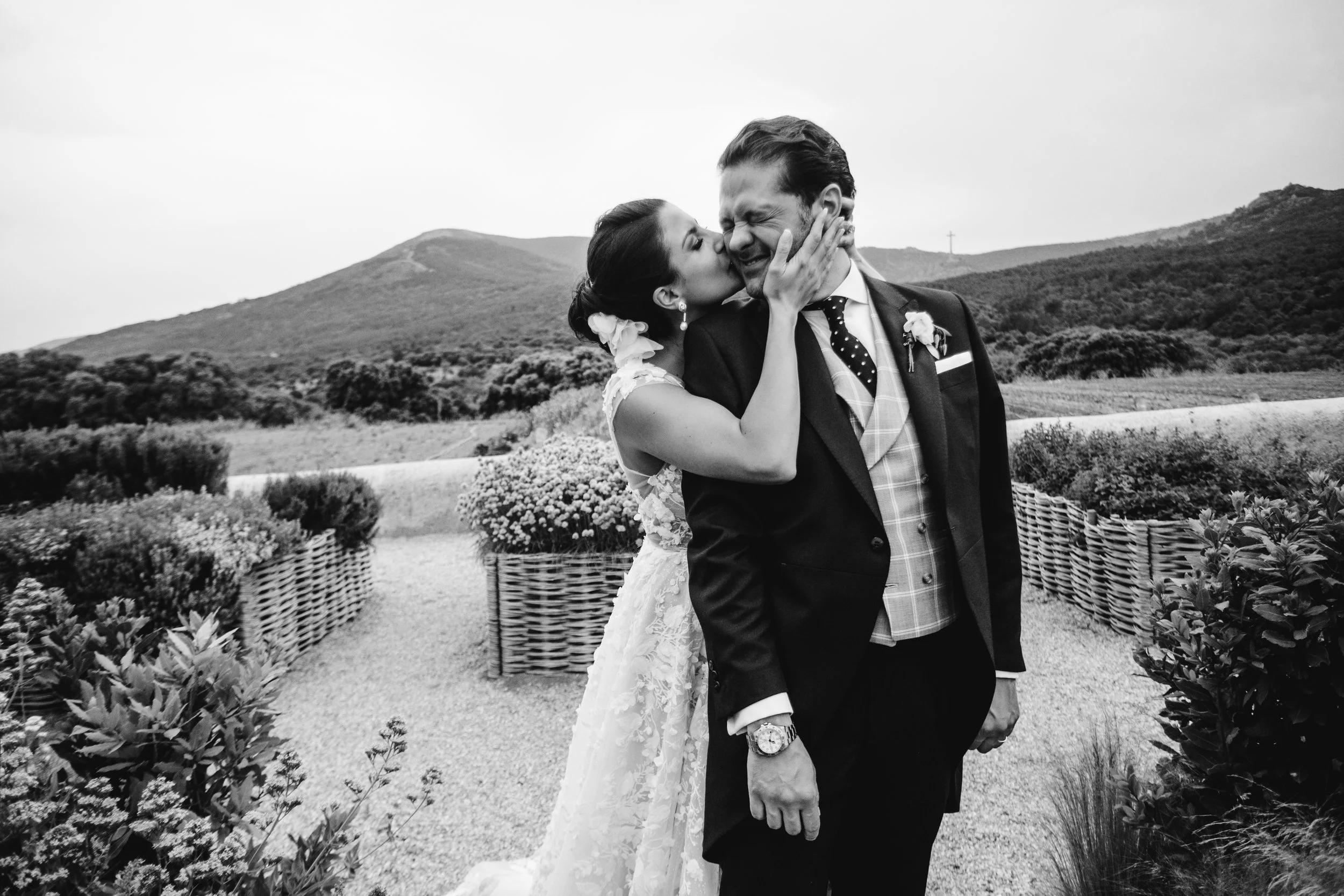 Una pareja de novios en una boda, la mujer le da un beso en la mejilla al hombre en un paisaje montañoso.