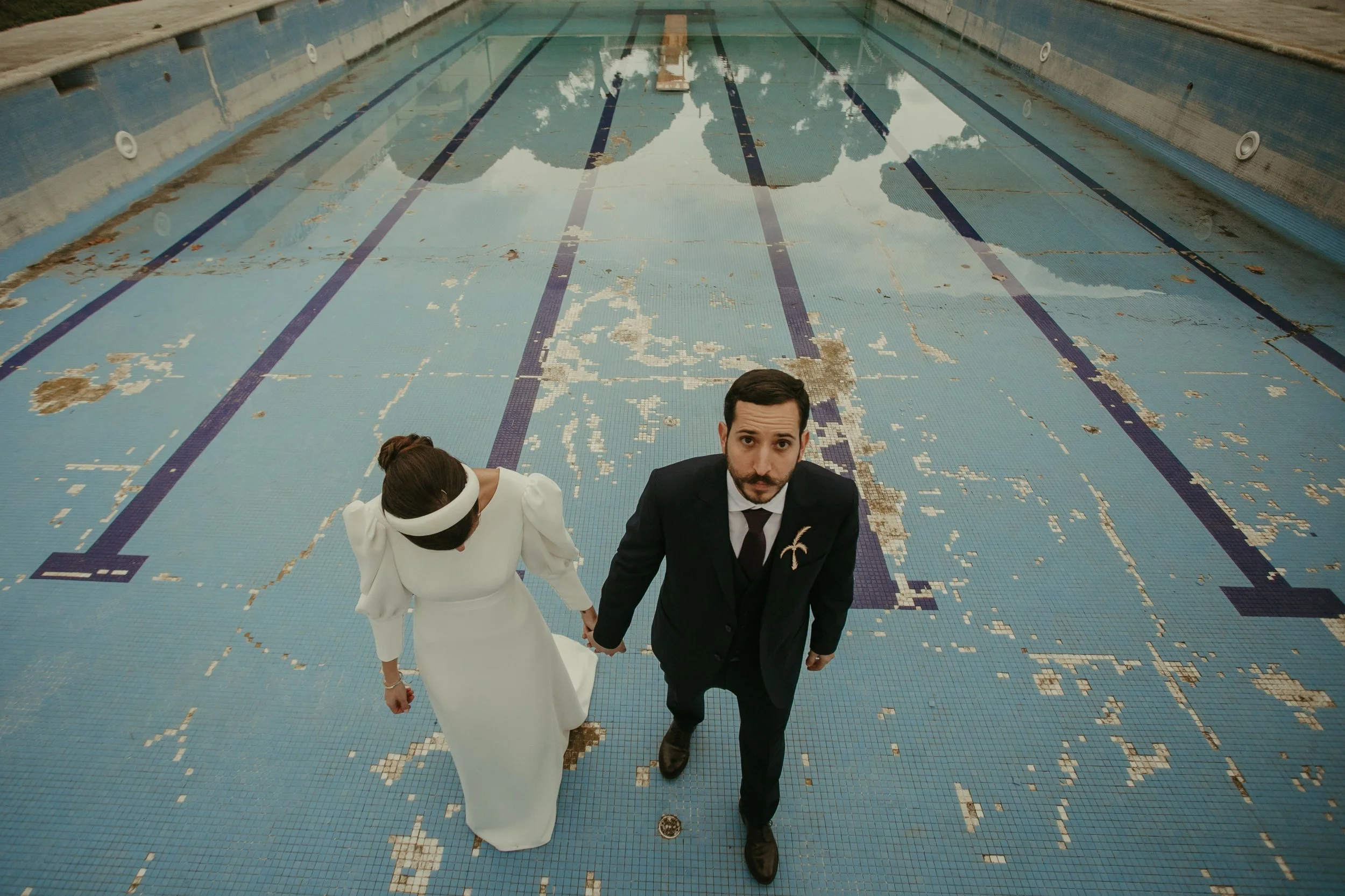 Pareja vestida de novios caminando en una piscina vacía y en mal estado, vista desde arriba.