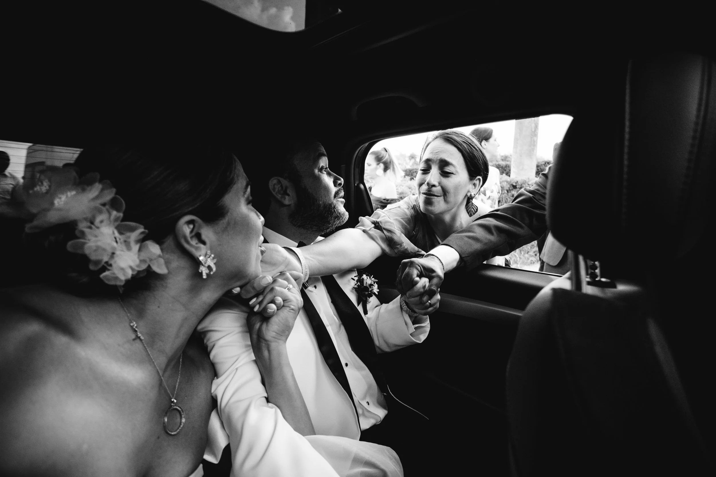 Una escena emotiva en la parte trasera de un vehículo con una pareja de boda y una mujer que les toca la mano desde el exterior, en un ambiente de celebración y alegría.