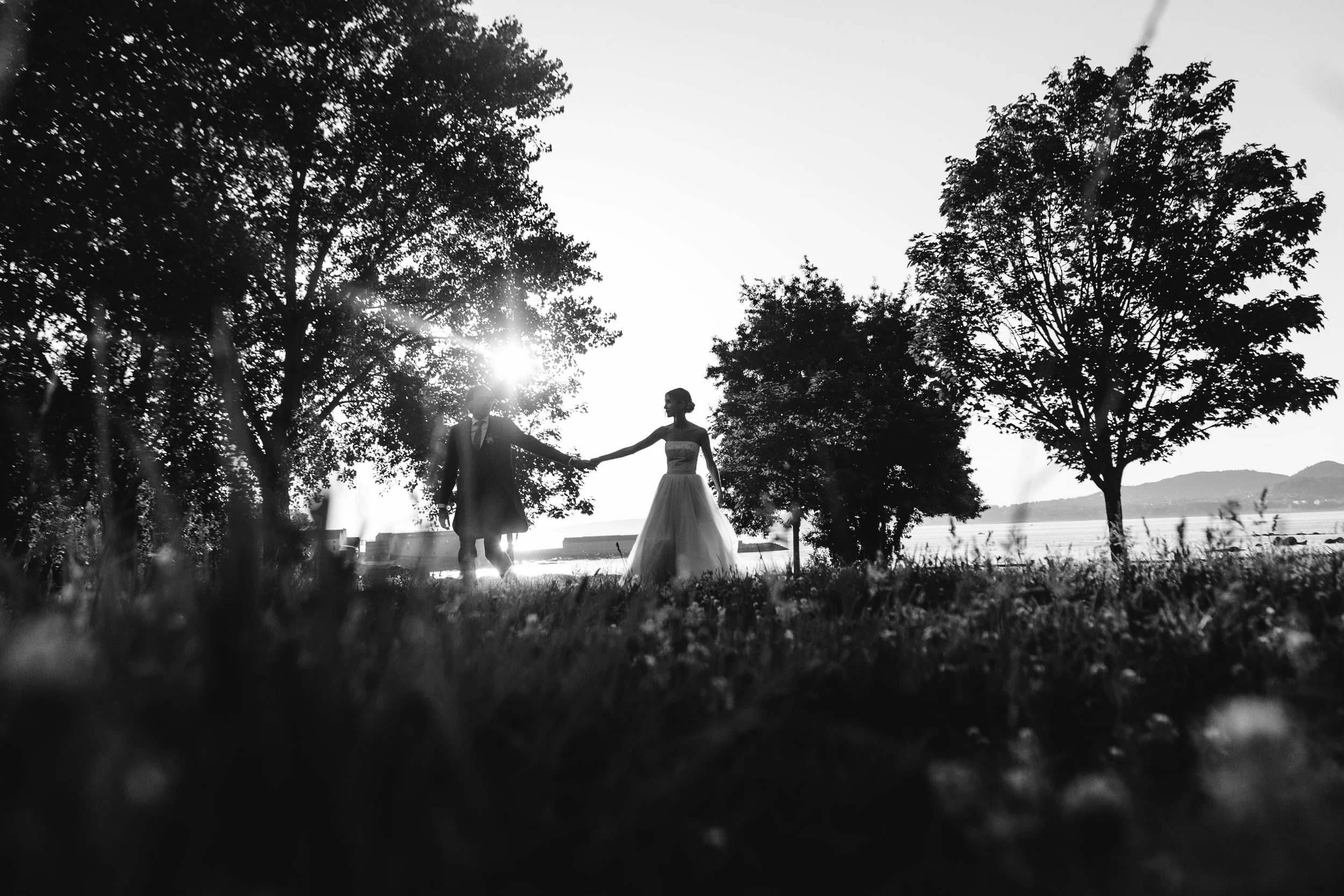 Pareja de novios en un escenario al aire libre, en un día soleado, en tonos en blanco y negro, sujetándose de las manos con árboles y un cuerpo de agua de fondo.