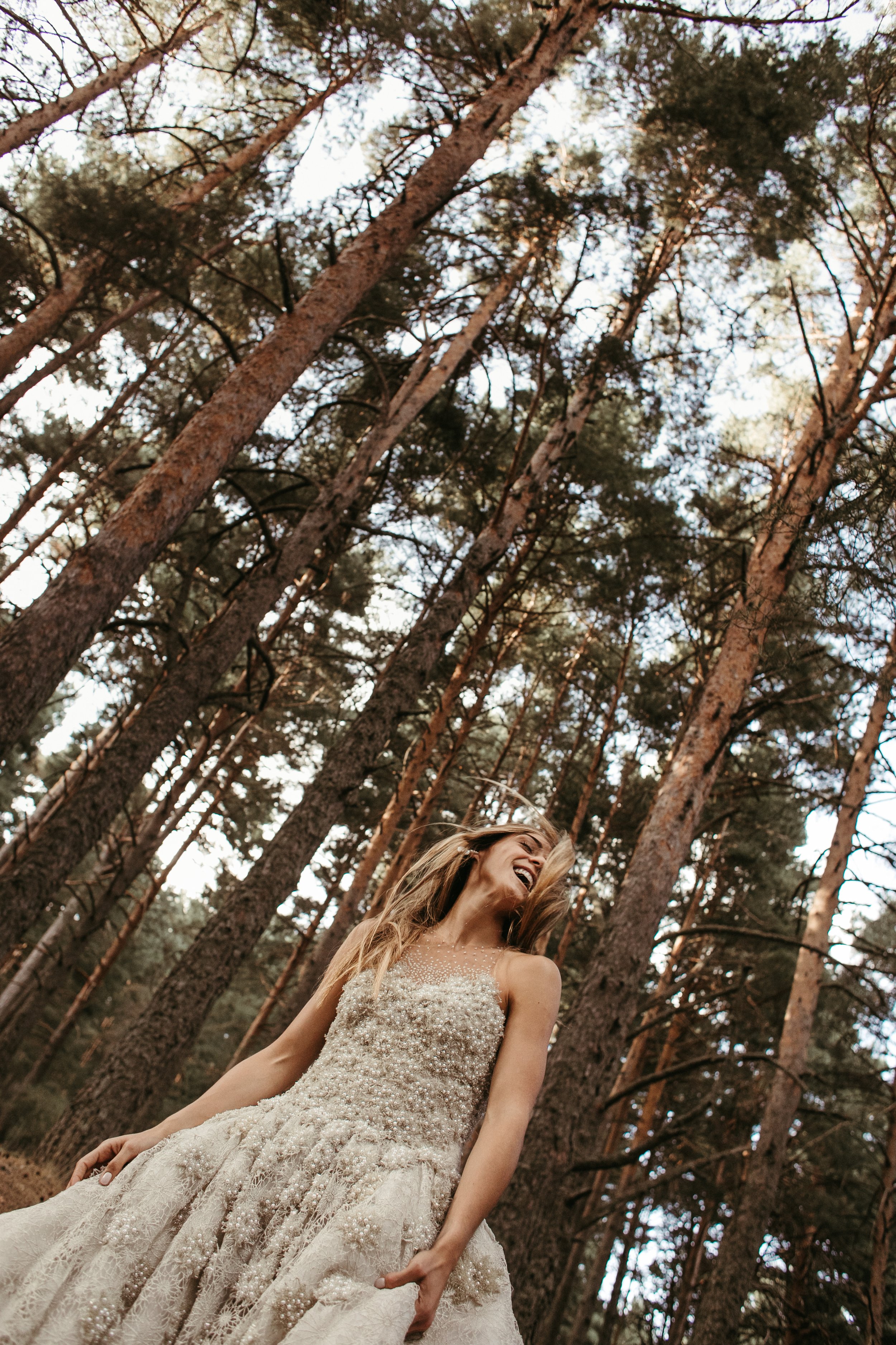 Una mujer sonriendo con un vestido de novia en un bosque con árboles altos y hojas verdes.