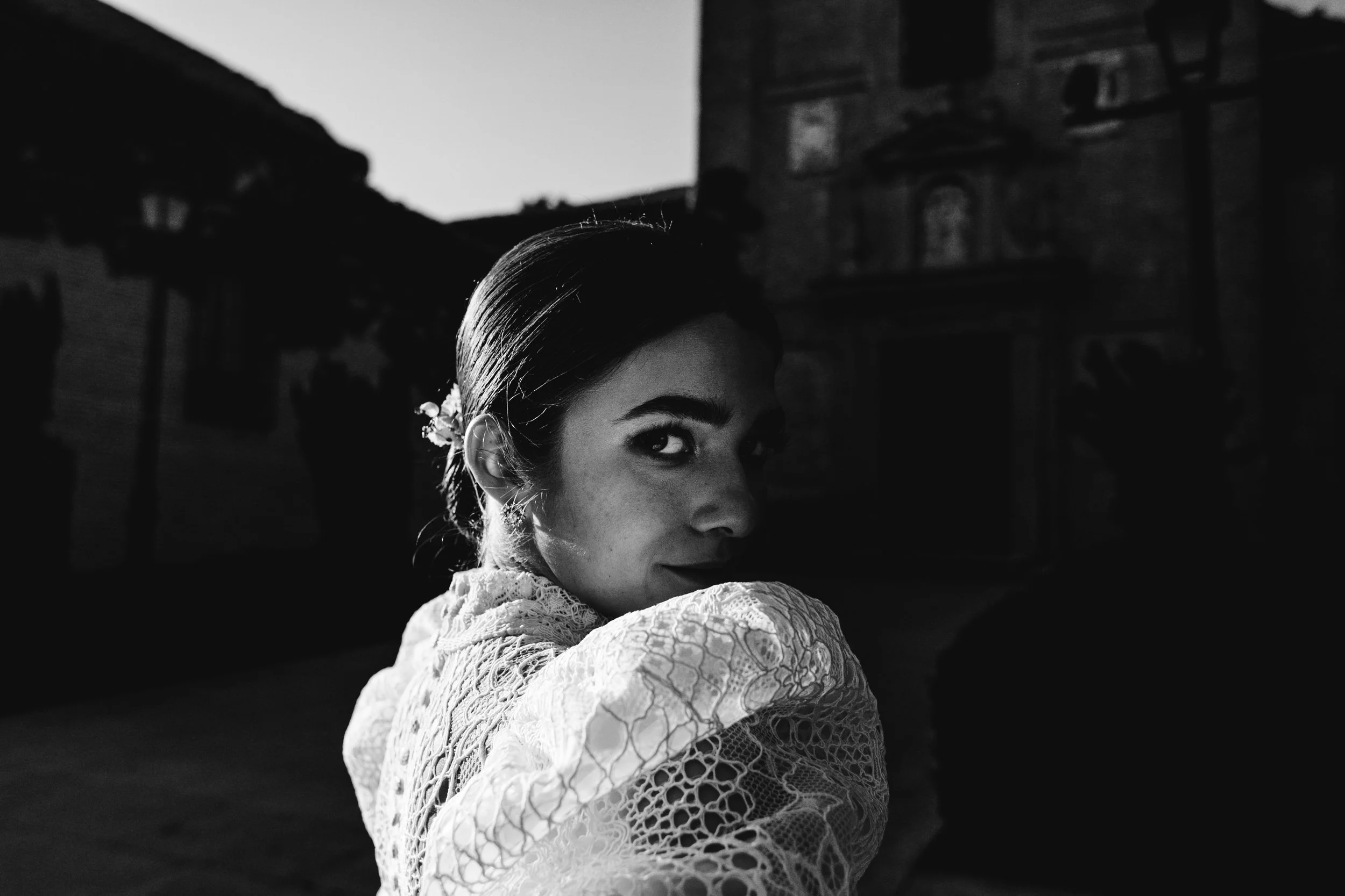 Retrato en blanco y negro de una mujer con cabello oscuro peinado hacia atrás, luciendo una expresión enigmática, en un entorno urbano con edificios antiguos de fondo durante la noche.