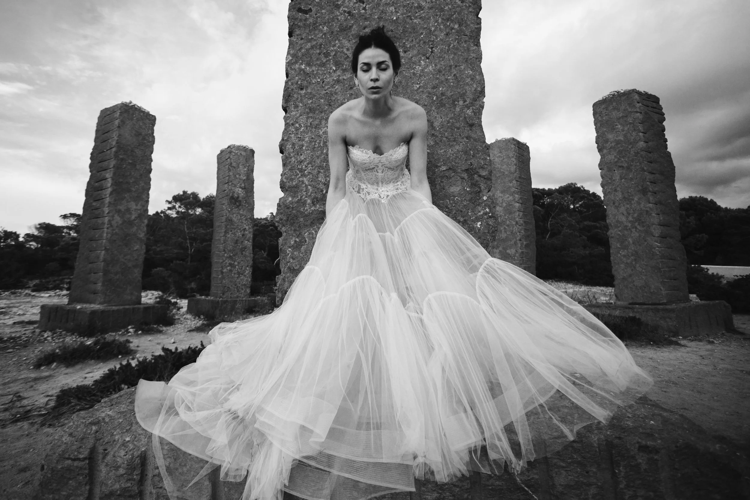 Mujer con vestido de novia blanco, sentada en una estructura de piedra antigua, al aire libre, en un día nublado, en una fotografía en blanco y negro.