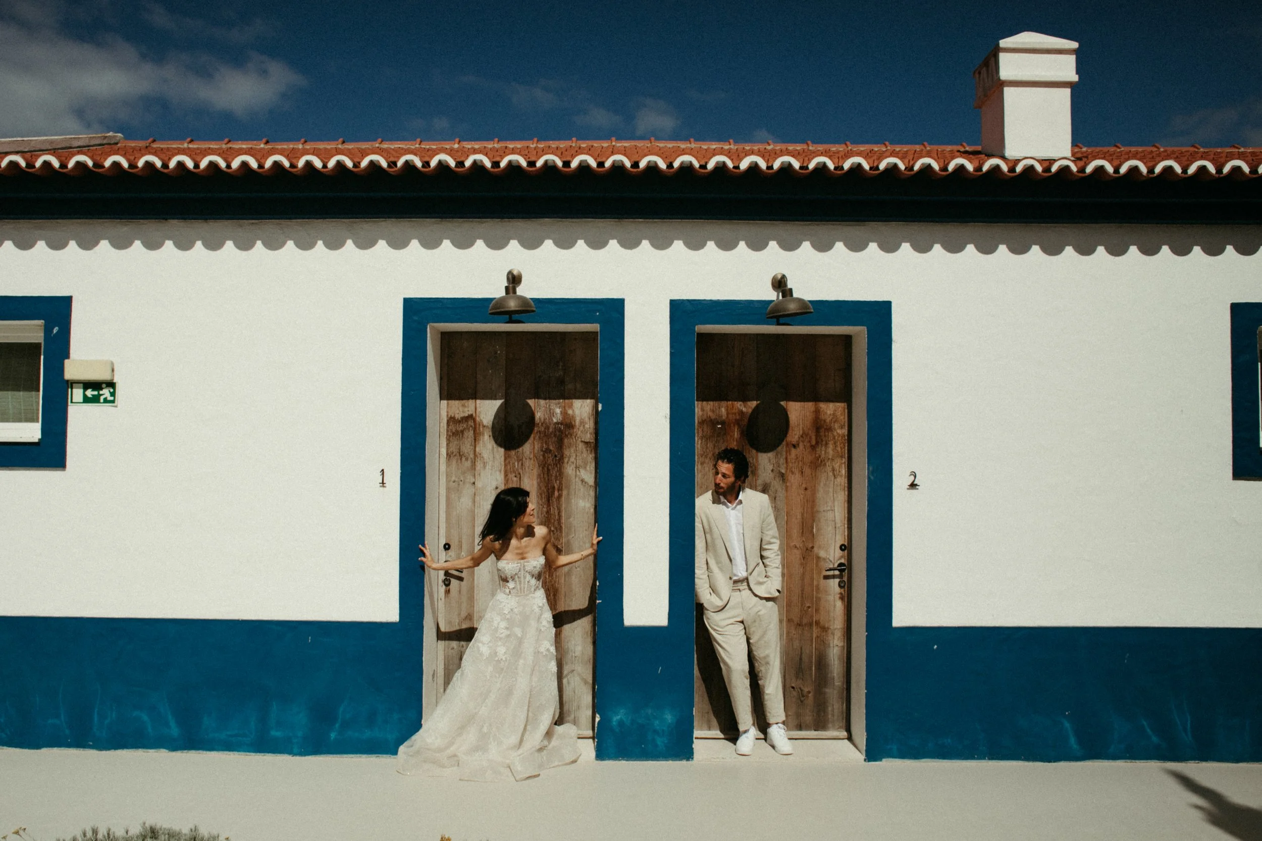 Pareja en trajes de boda posando frente a puertas de madera en una casa blanca con acento azul y techo de tejas rojas bajo cielo azul.