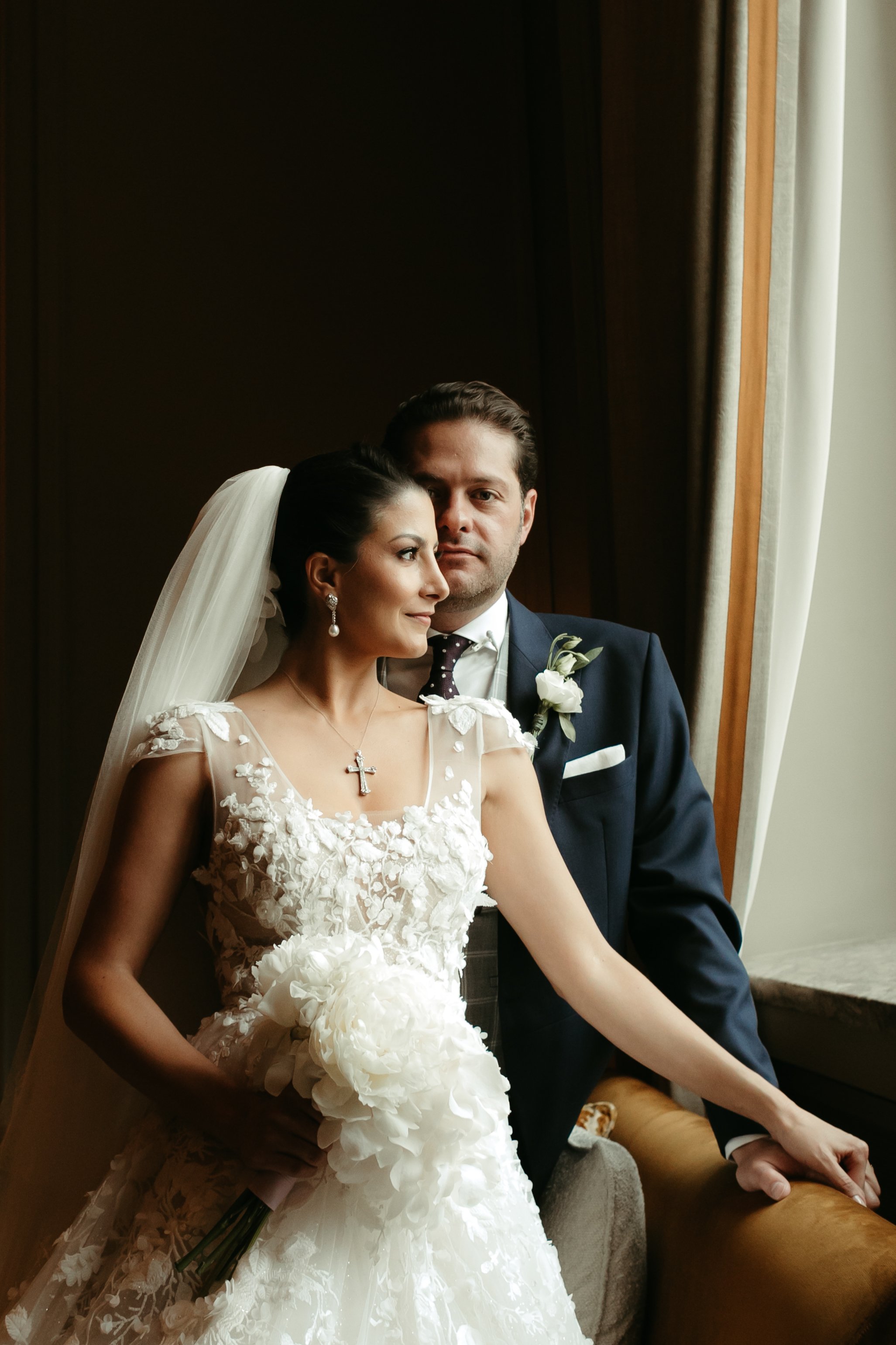 Pareja de novios en boda, la novia con vestido de encaje blanco y velo, sosteniendo ramos de flores blancas, el novio con traje oscuro y corbata con puntos blancos, mirando por una ventana