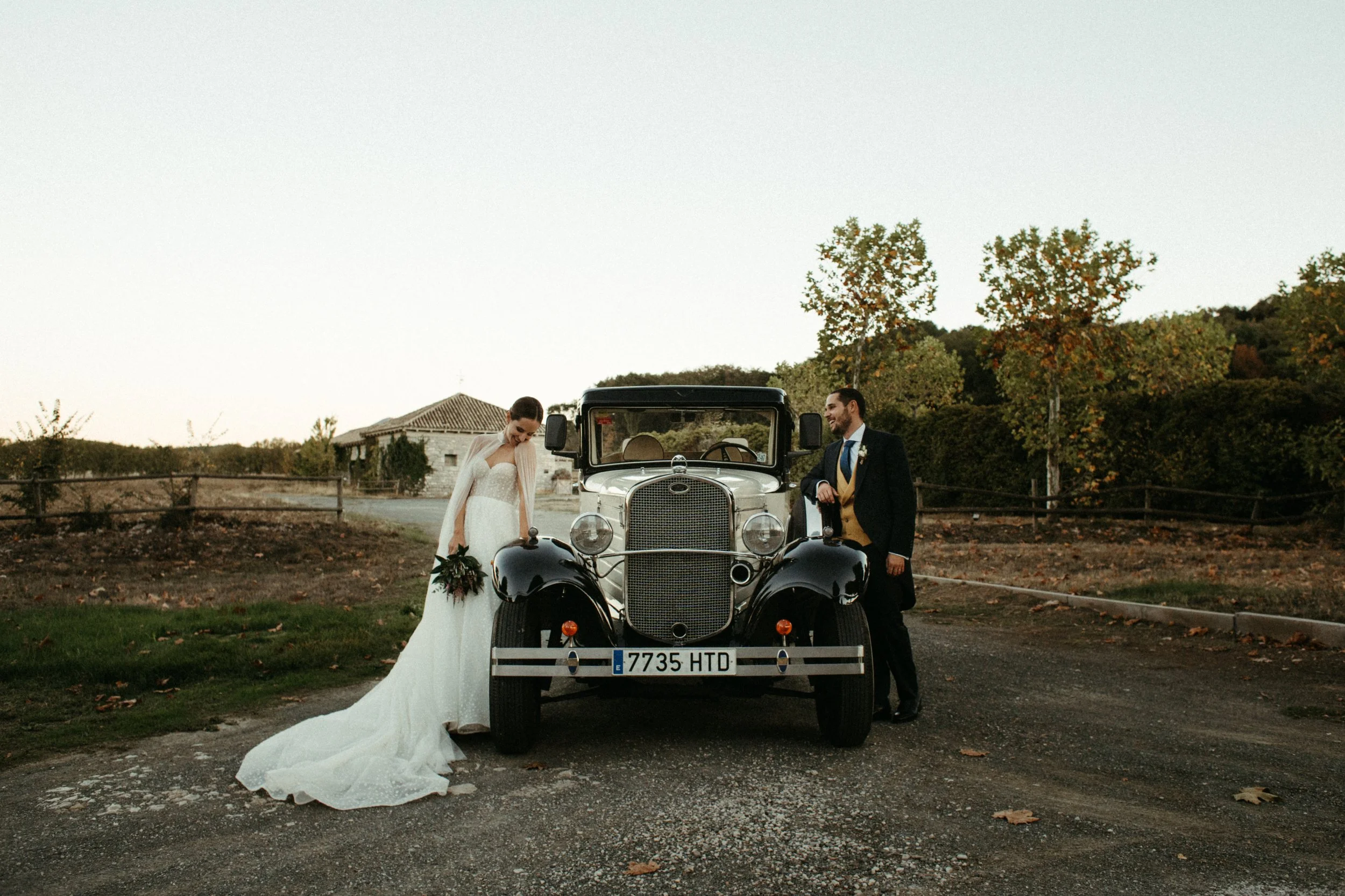 Una pareja de novios en su boda, posando junto a un auto antiguo en un entorno rural con árboles y una casa en el fondo.