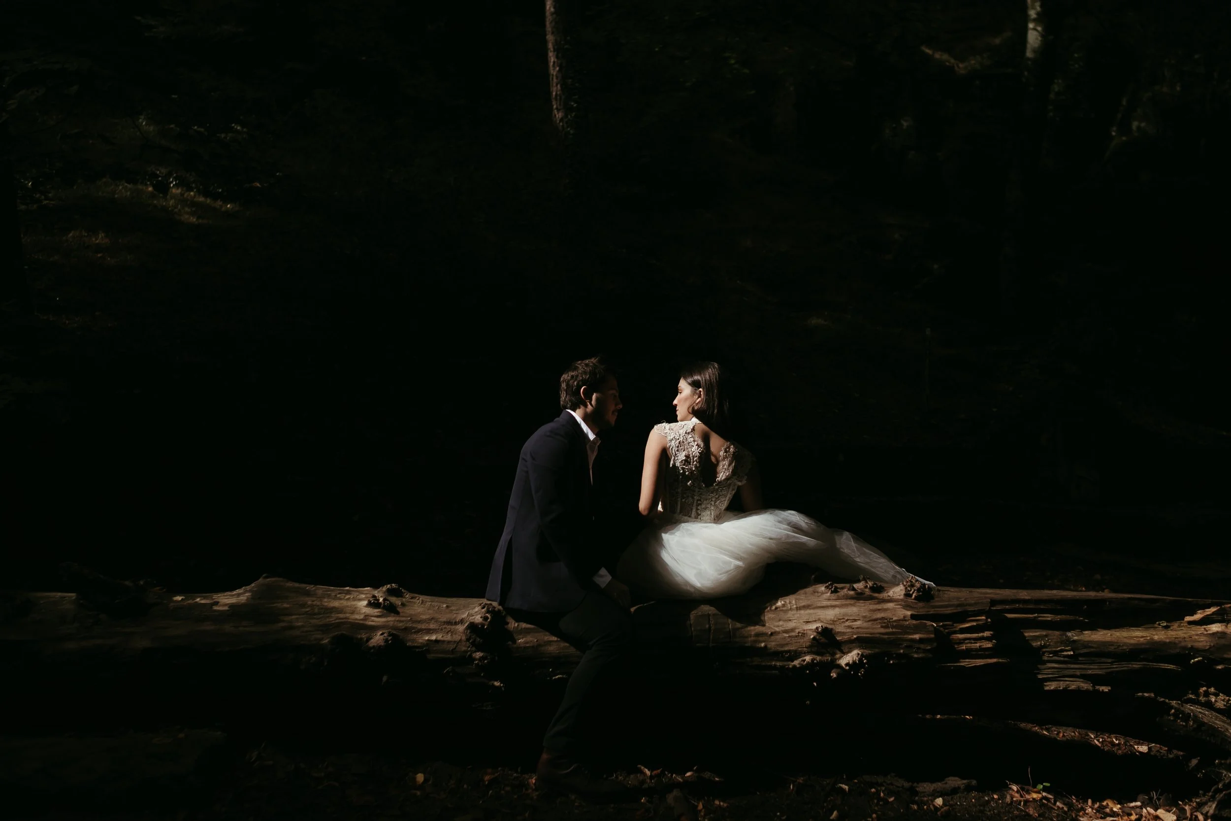 Pareja de novios en un entorno oscuro, sentados en un tronco, con iluminación enfocada en ellos.
