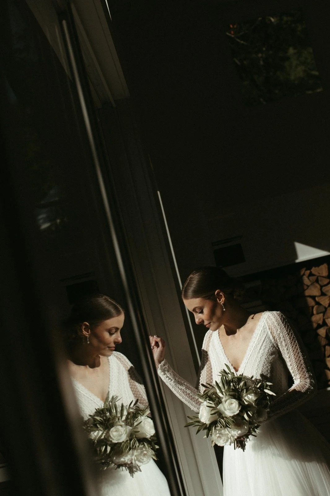 Mujer vestida de novia en reflejo en un espejo, sosteniendo un ramo de flores blancas y mirando hacia abajo, en un ambiente interior con lámpara