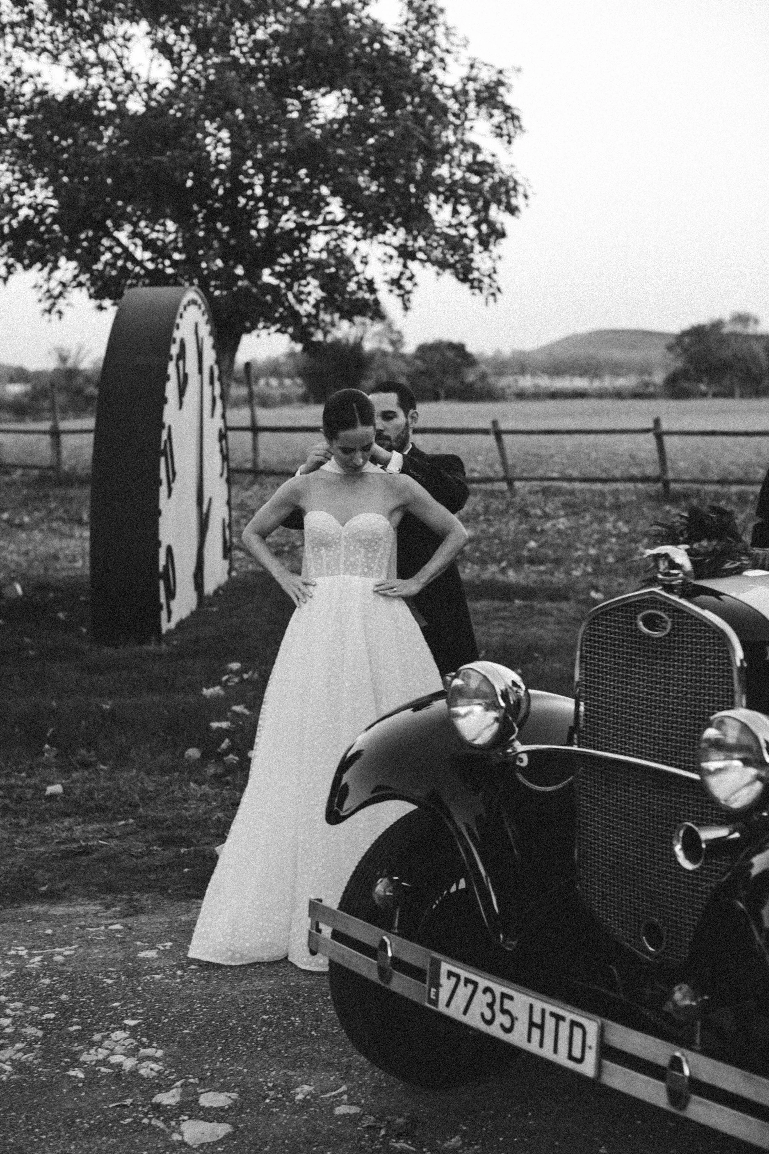 Pareja de novios en una sesión de fotos, la novia con vestido de novia y cabello recogido, el novio de traje oscuro, en un paisaje al aire libre con árboles y una cerca de madera.