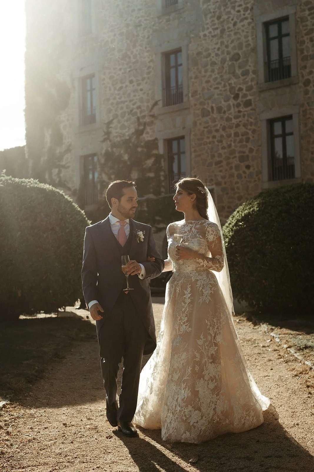 Bride and groom walking in golden afternoon light beside an old stone building on their wedding day in El Escorial, Madrid