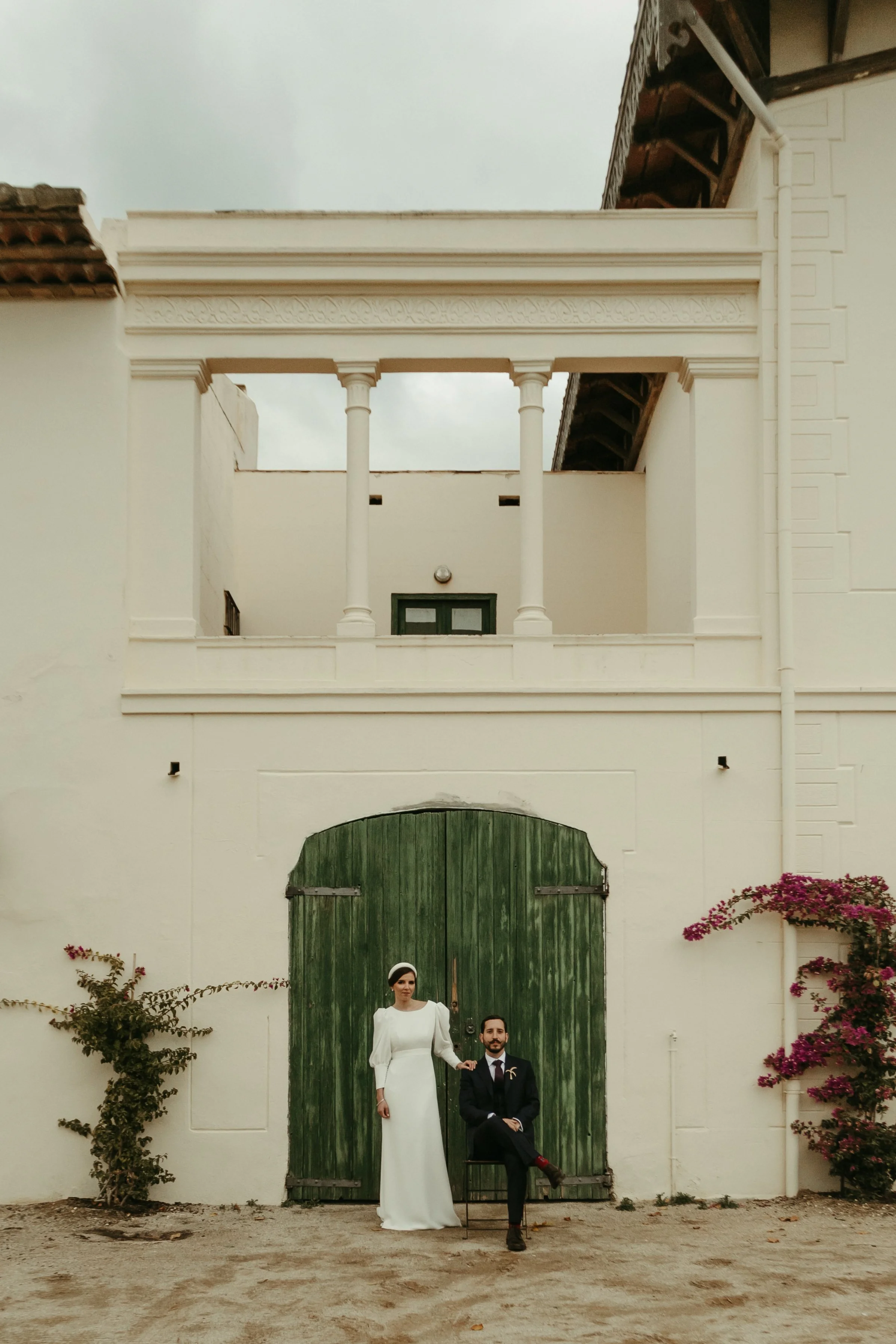 Pareja de recién casados con vestimenta formal posando frente a una porta de madera verde en un edificio de color blanco, con plantas de flores a los lados.