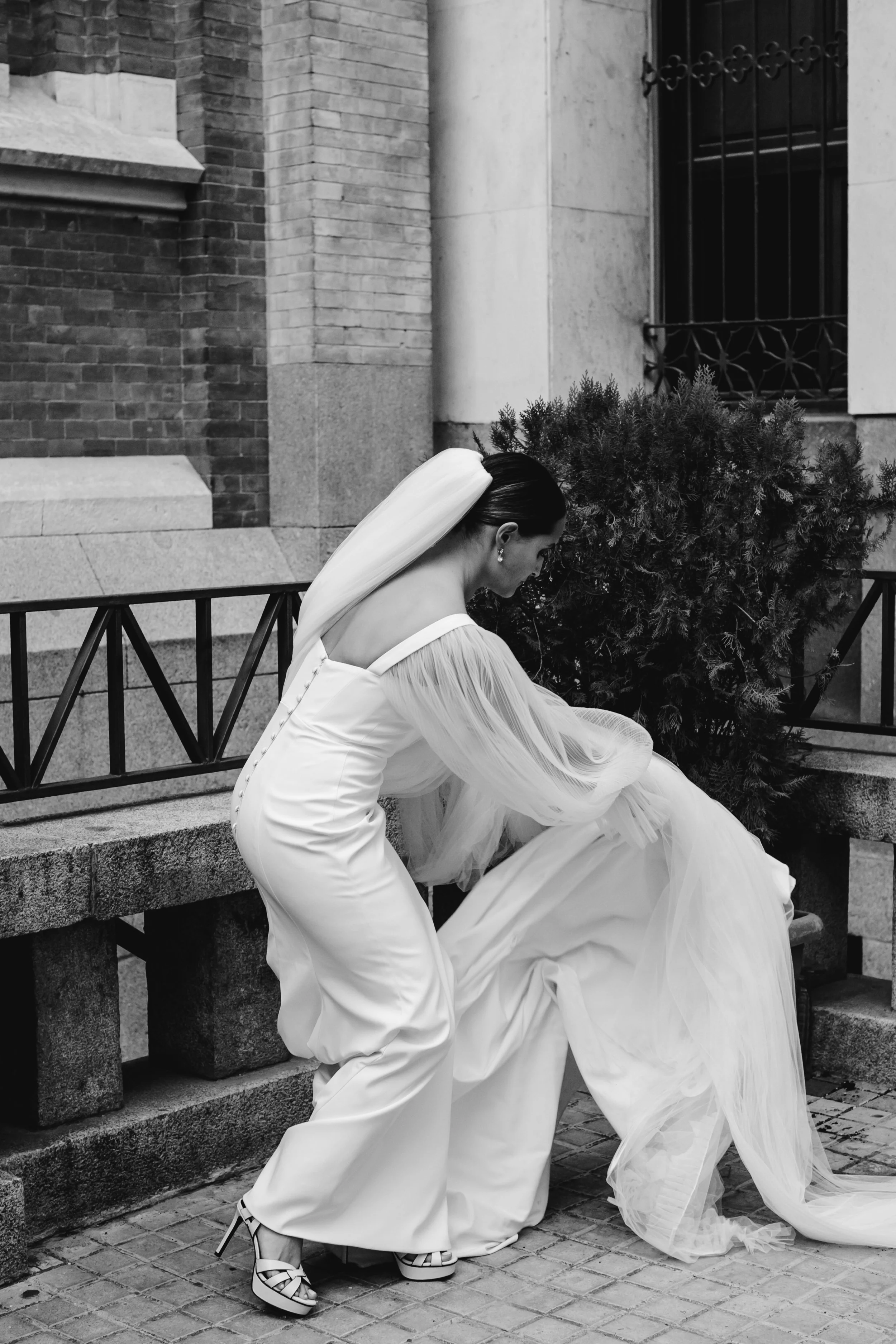 Mujer con vestido de novia ajustándose en una banca de piedra en la calle, en un entorno urbano
