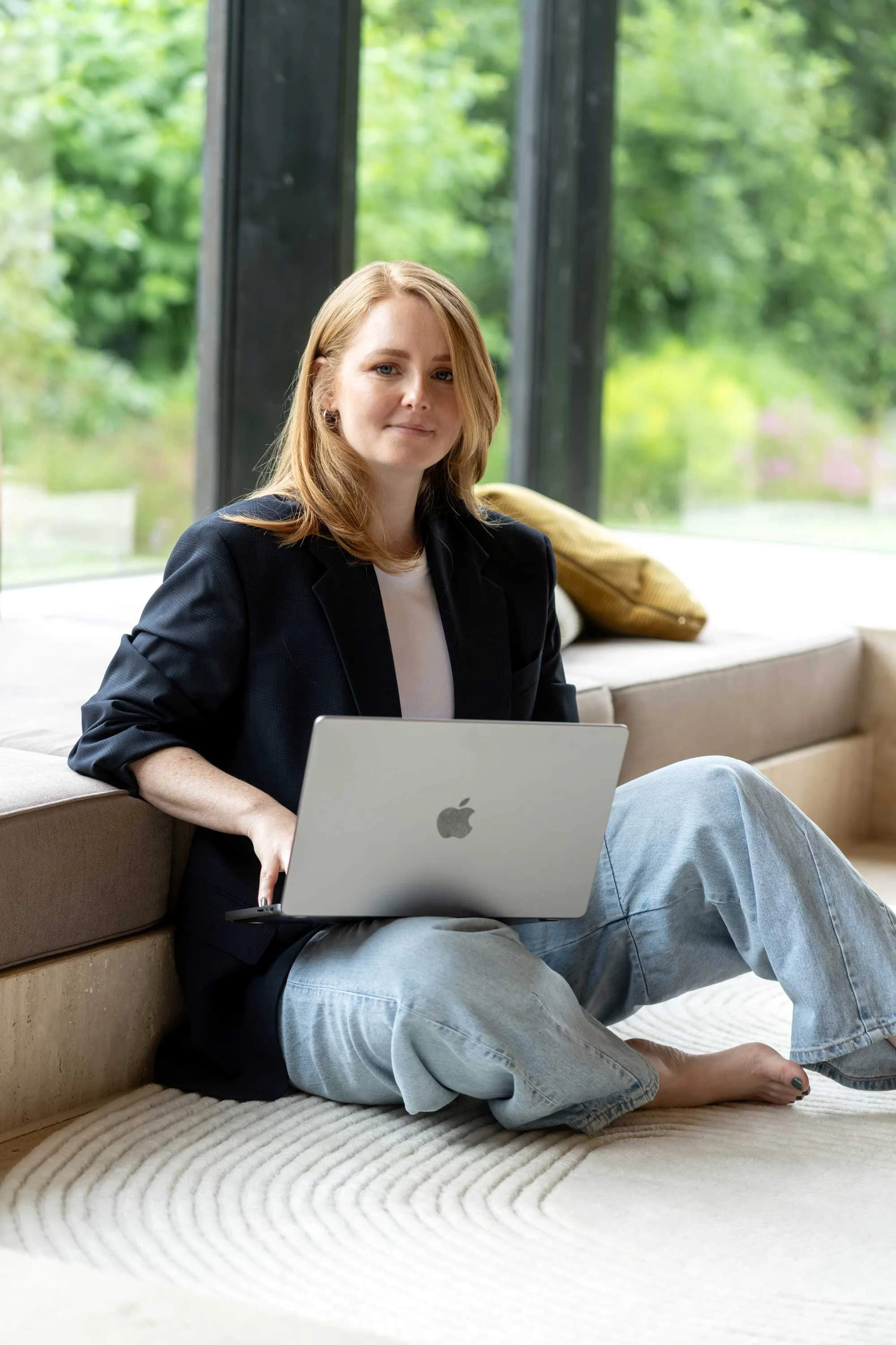 Junge Frau mit Laptop, sitzend auf dem Boden vor großen Fenstern mit grüner Aussicht, im Wohnzimmer.