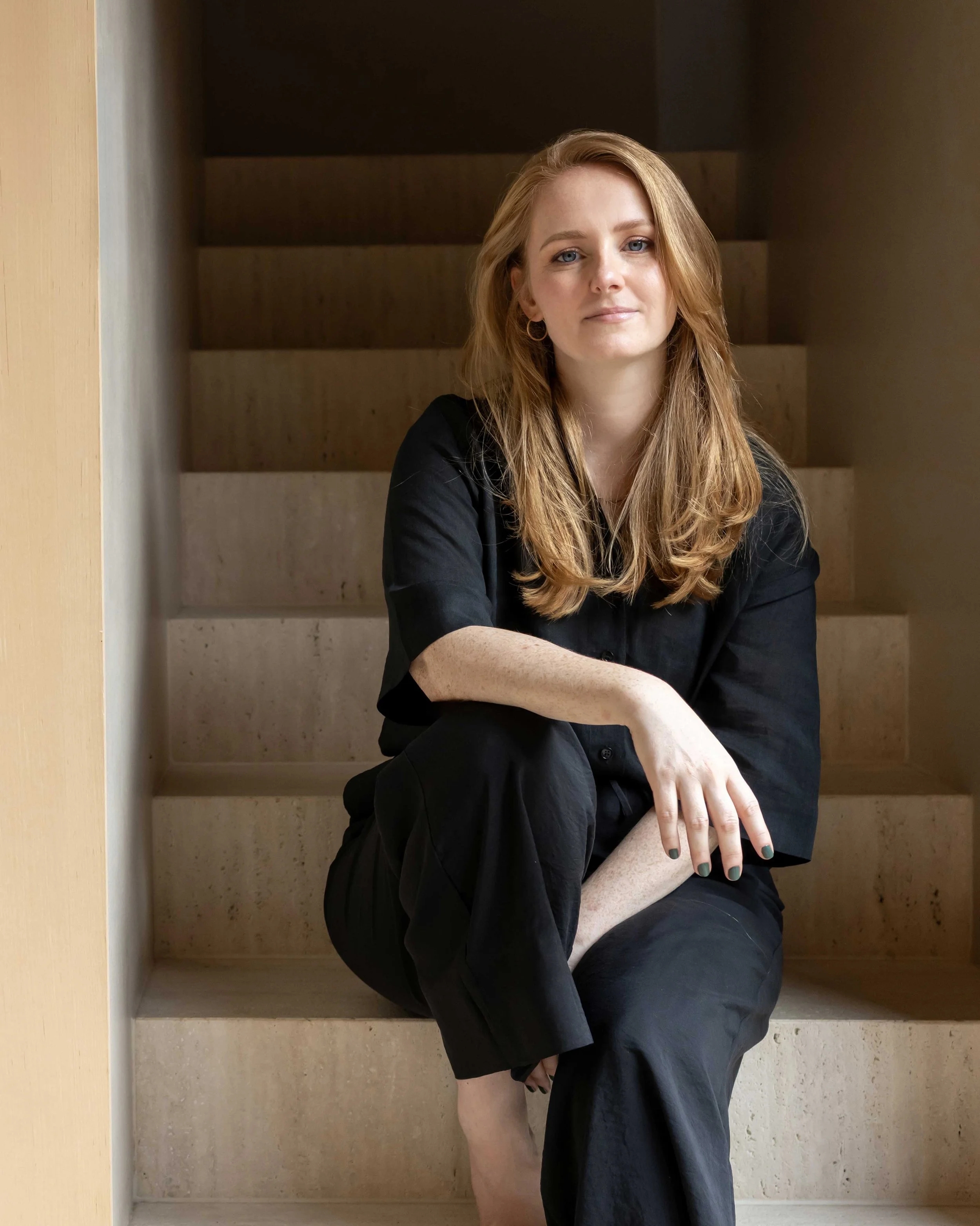 Eine Frau mit langen roten Haaren sitzt entspannt auf einer Treppe, trägt eine schwarze Kleidung und blickt in die Kamera.