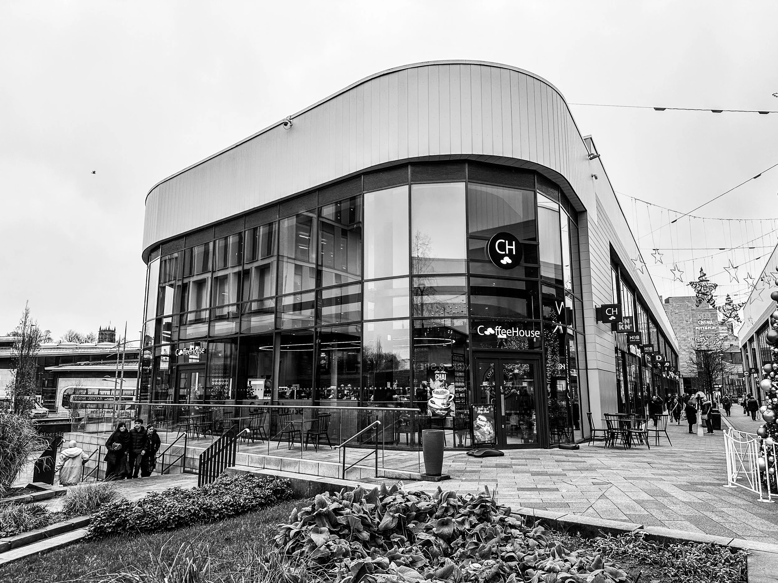 A modern shopping mall with a café on the ground floor, glass windows, and outdoor seating, with pedestrians walking on the street decorated with Christmas ornaments.