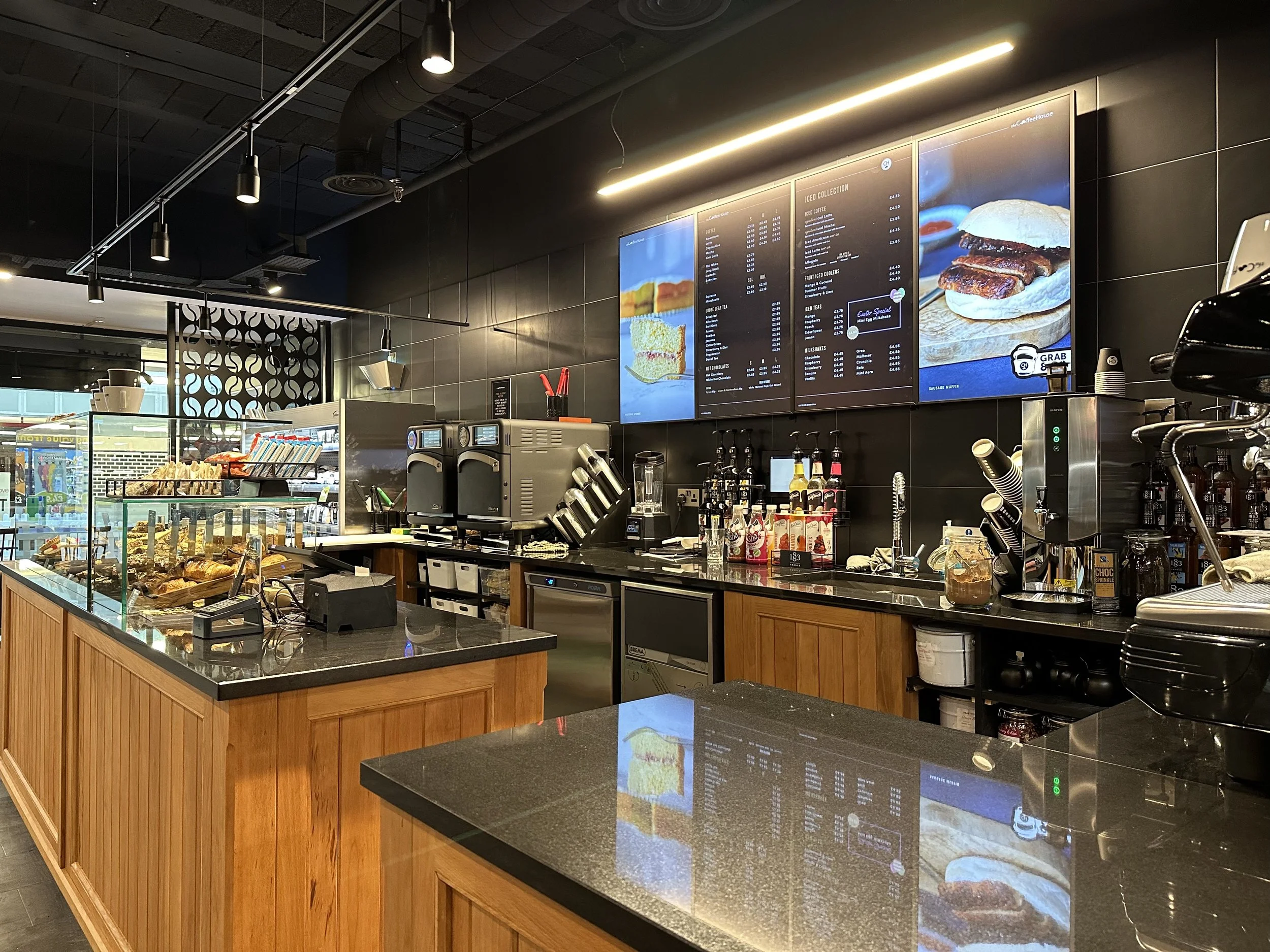 Modern coffee shop interior with wooden counters, pastries displayed in glass cases, and menu boards on the wall. Coffee machines and syrup bottles are visible behind the counter.