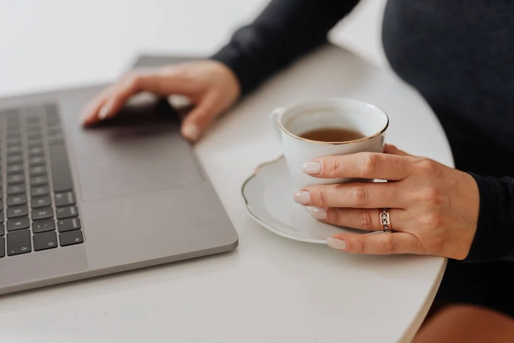 Person holding a cup of tea or coffee with a silver ring on finger, working on a laptop at a white table.