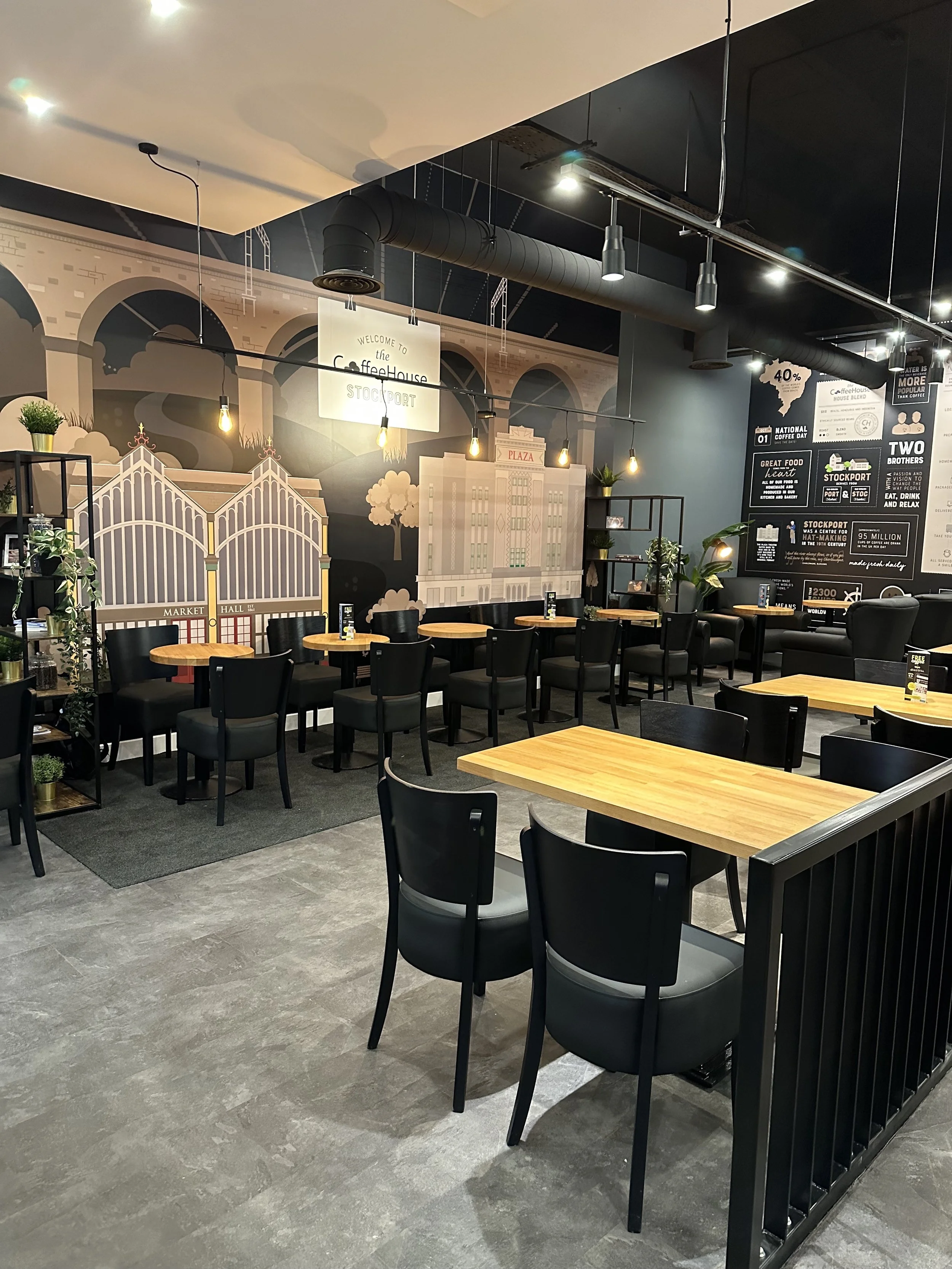 Interior of a modern coffee shop with wooden tables and black chairs, featuring a decorative mural, pendant lighting, and black ceiling ducts. Indoor plants and shelving add decor.
