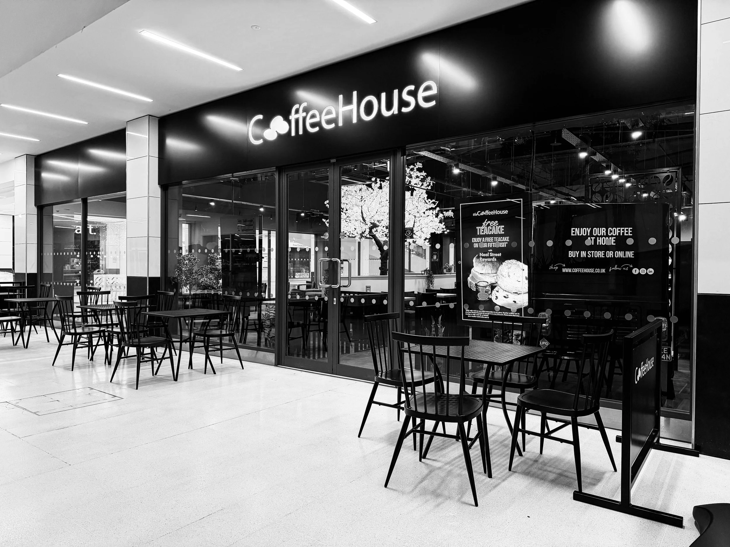 Black and white photo of a CoffeeHouse café storefront with outdoor seating, including tables and chairs, inside a shopping mall.