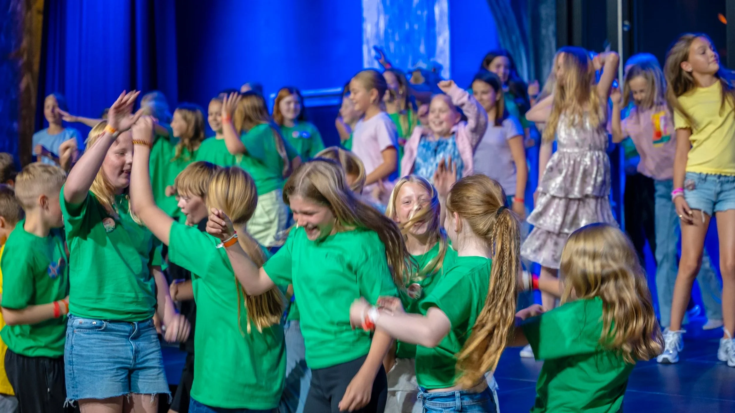 Flere barn ikledd fargerike klær. De synger på en scene. Fremst på raden står flere jenter i grønne t-skjorter som ser ut som de danser.