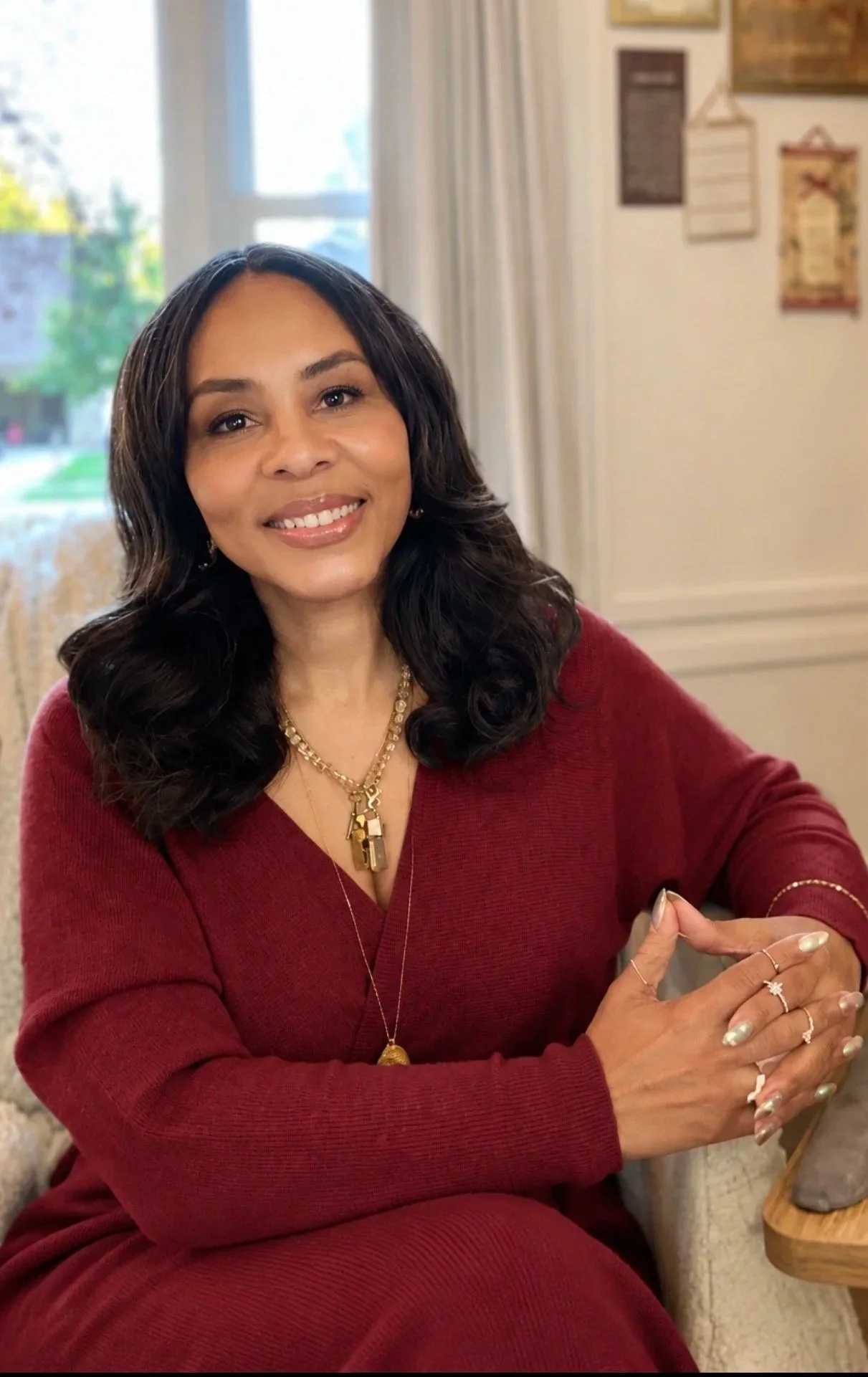 Misha Williams smiling in a burgundy sweater, seated with her hands clasped in a sunlit room